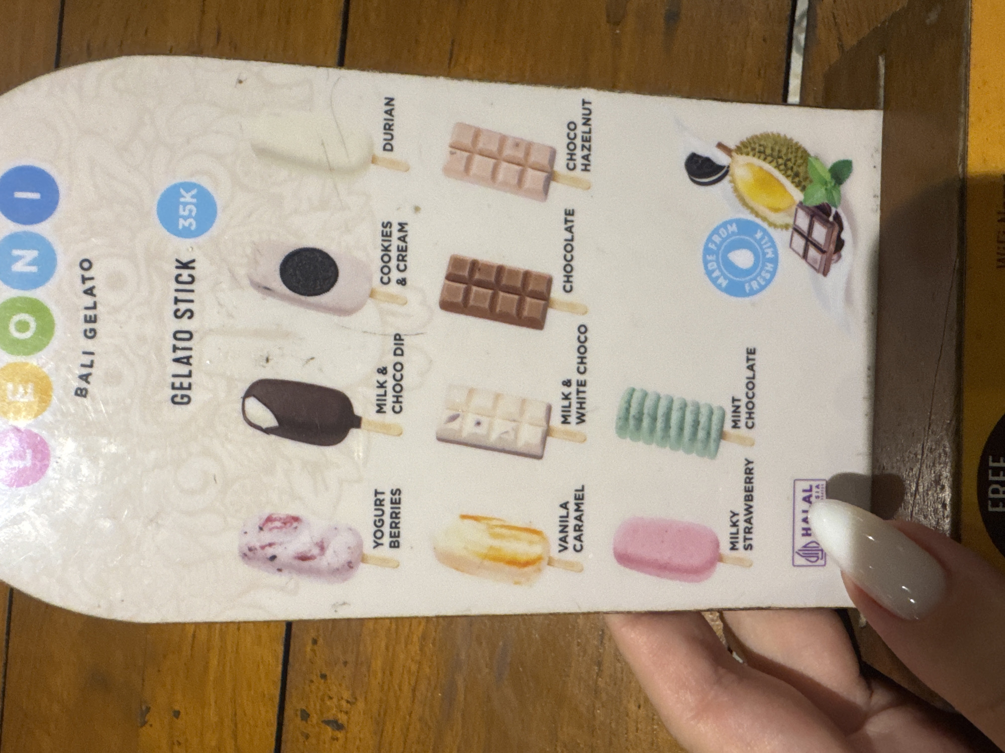 The image shows a person holding a gelato stick menu from "INONI BALI GELATO" on a wooden table. The scene appears to be indoors, likely in a cafe or restaurant in Nusa Penida, Indonesia. The menu is oriented sideways and features illustrations of various gelato sticks.
The prominent text "GELATO STICK 35K" indicates a price of 35,000 (presumably Indonesian Rupiah) per stick. Ten different flavors are listed: DURIAN, CHOCO HAZELNUT, COOKIES & CREAM, CHOCOLATE, MILK & CHOCO DIP, MILK & WHITE CHOCO, YOGURT BERRIES, VANILA CARAMEL, MILKY STRAWBERRY, and MINT CHOCOLATE.
Notable details include a circular logo stating "MADE FROM FRESH MILK" and a "HALAL SIA" certification mark. The person's hand has polished, light-colored nails, holding the menu against a dark brown wooden plank table. A partially visible yellow strip on the right edge also shows the word "FREE."