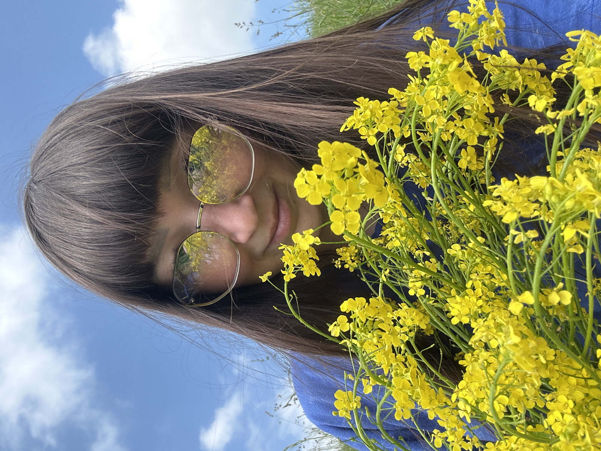 A medium shot captures a young woman from the chest up, holding a bouquet of bright yellow flowers in front of her. She is wearing a royal blue top and gold-rimmed glasses. Her long, dark brown hair with bangs partially covers her forehead and the top of her glasses. The glasses' lenses reflect the blue sky with scattered white clouds and more yellow flowers, suggesting an outdoor setting. The background consists of a vibrant blue sky with wispy clouds and out-of-focus green grass and plants, indicating a natural, possibly rural, environment. The scene is set in or near the village of Sredneye Kechevo, Russia, evident from the visible flora characteristic of the region.