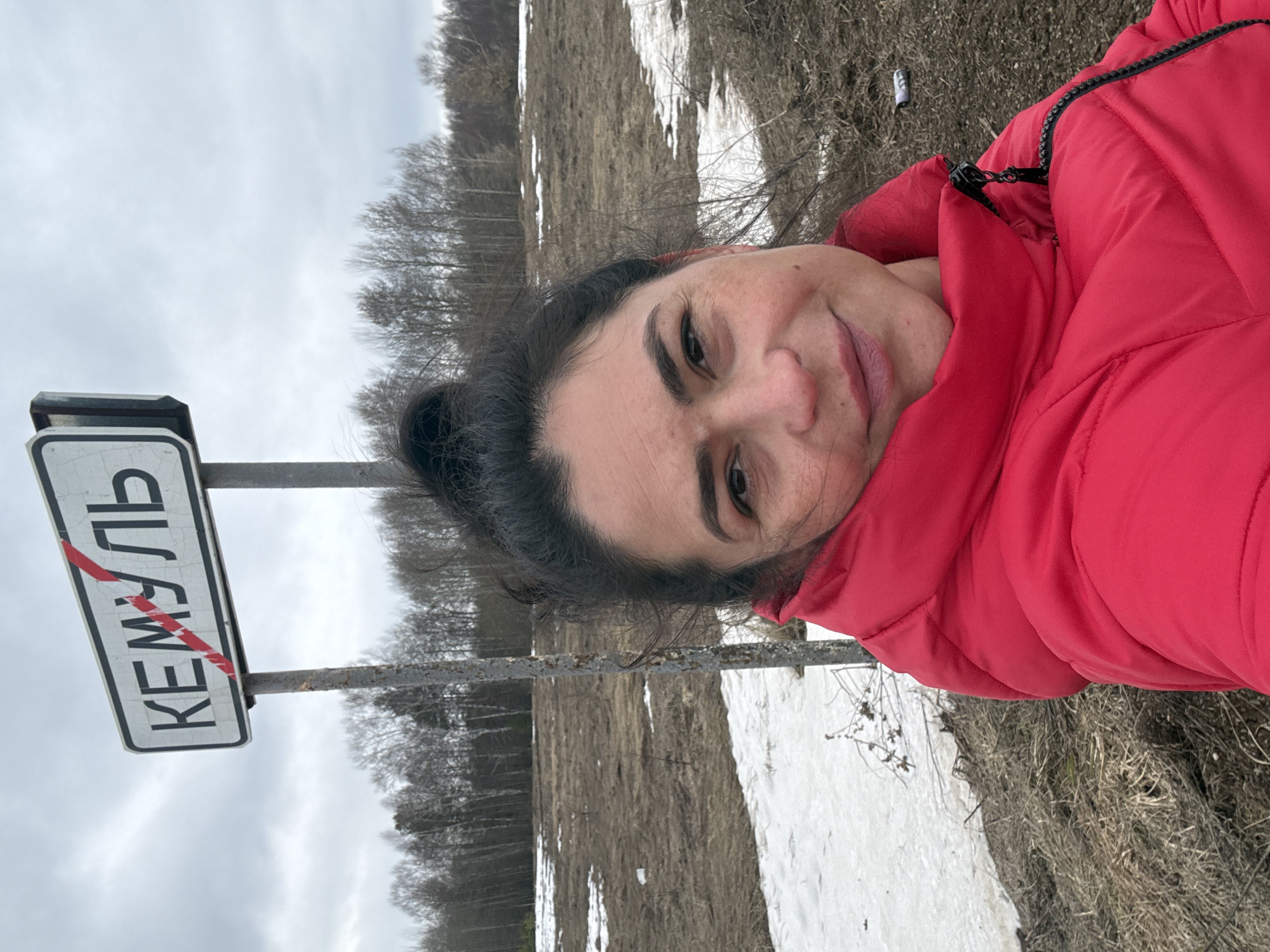 A woman in a red jacket stands in front of a sign indicating the city of Kemul, Russia. The sign is white with black lettering, and the city name "КЕМУЛЬ" is written in Cyrillic script. A red line is drawn across the sign, indicating that the city has been crossed out. The background consists of a field with some snow patches, trees, and an overcast sky.