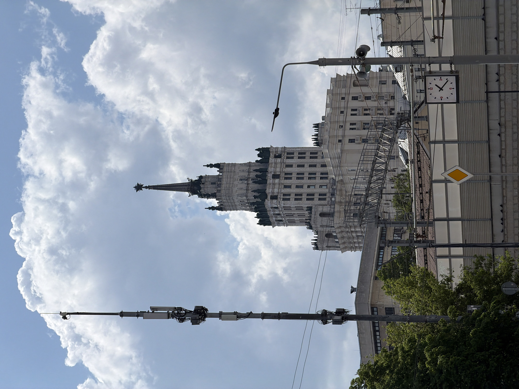 The image shows a view in Moscow, Russia, featuring the Kotelnicheskaya Embankment Building, one of the "Seven Sisters" Stalinist skyscrapers. The building is prominent against a cloudy sky. Foreground elements include utility poles with attached equipment, a street lamp, a building wall with a clock displaying the time around 3:00 PM, and a "yield" road sign. A tree is visible at the bottom.