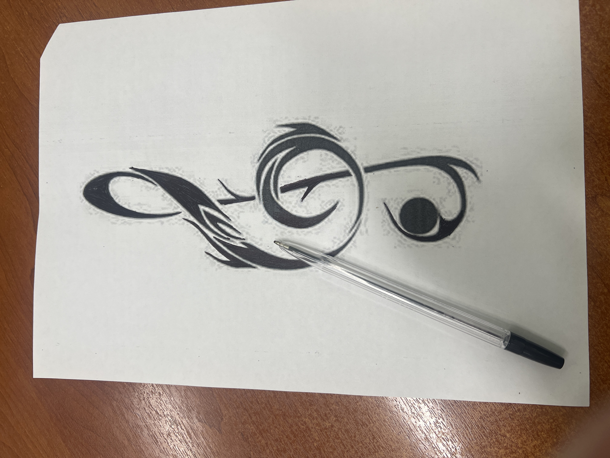 An overhead, slightly angled shot shows a sheet of white paper on a wooden surface, possibly a desk. The paper features a drawing of a treble clef, rendered in black with shading. A clear, plastic ballpoint pen with a black tip rests diagonally across the paper, its tip hovering just above the drawing. The wooden surface has a visible grain pattern.