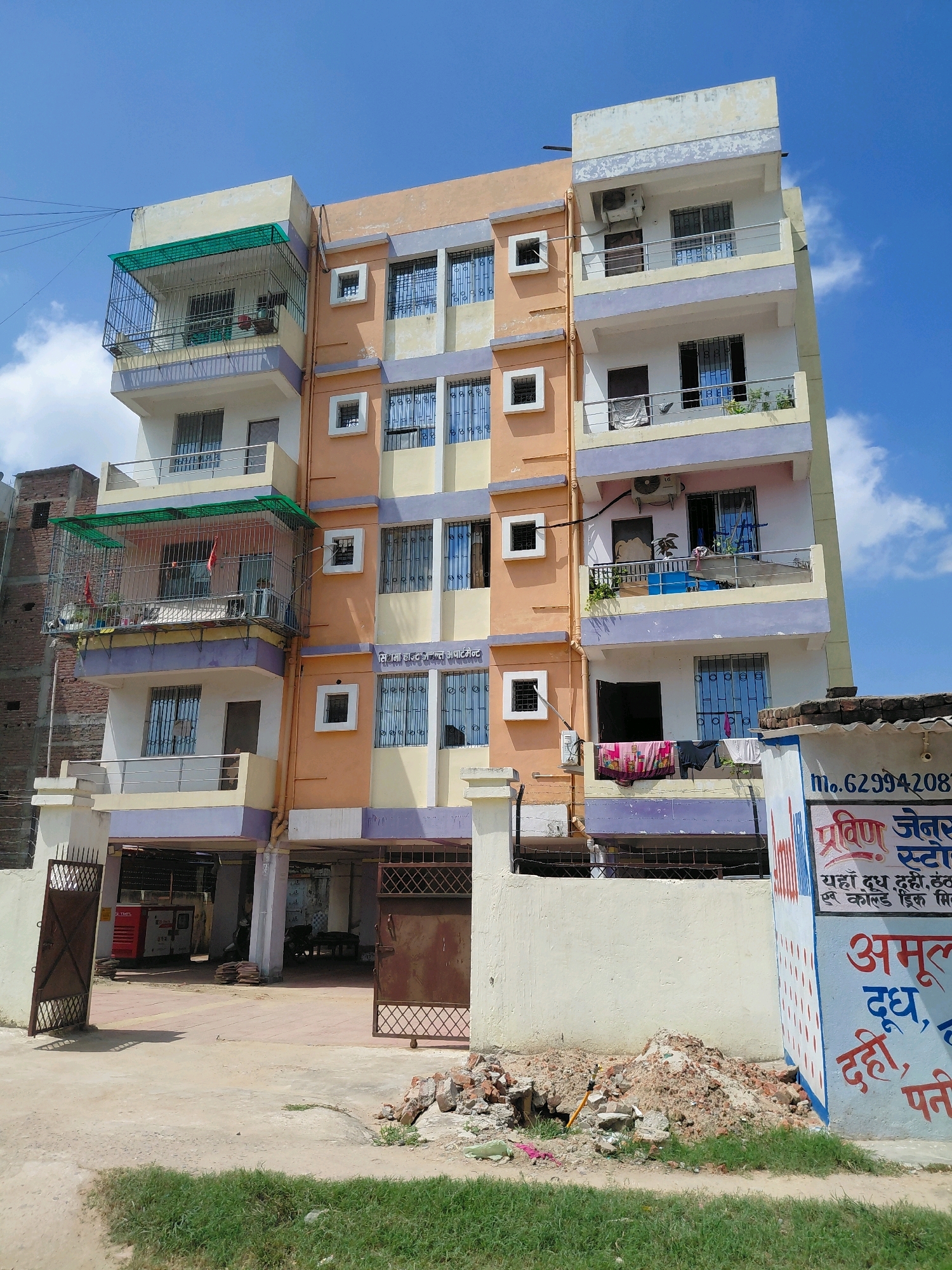 The image shows a multi-story apartment building with an orange and cream facade, situated on a street in Patna, India. The building has multiple balconies with metal railings and some are enclosed with metal grates. Several windows are visible, many with decorative metal grills. On one balcony, laundry is hung out to dry. In the foreground, there are open metal gates, a pile of construction debris, and green grass. A sign with Hindi text is partially visible on the right side of the image, with some text also visible on a blue and white structure next to it. The sky is blue with scattered clouds. The overall scene depicts a residential area in an urban setting.