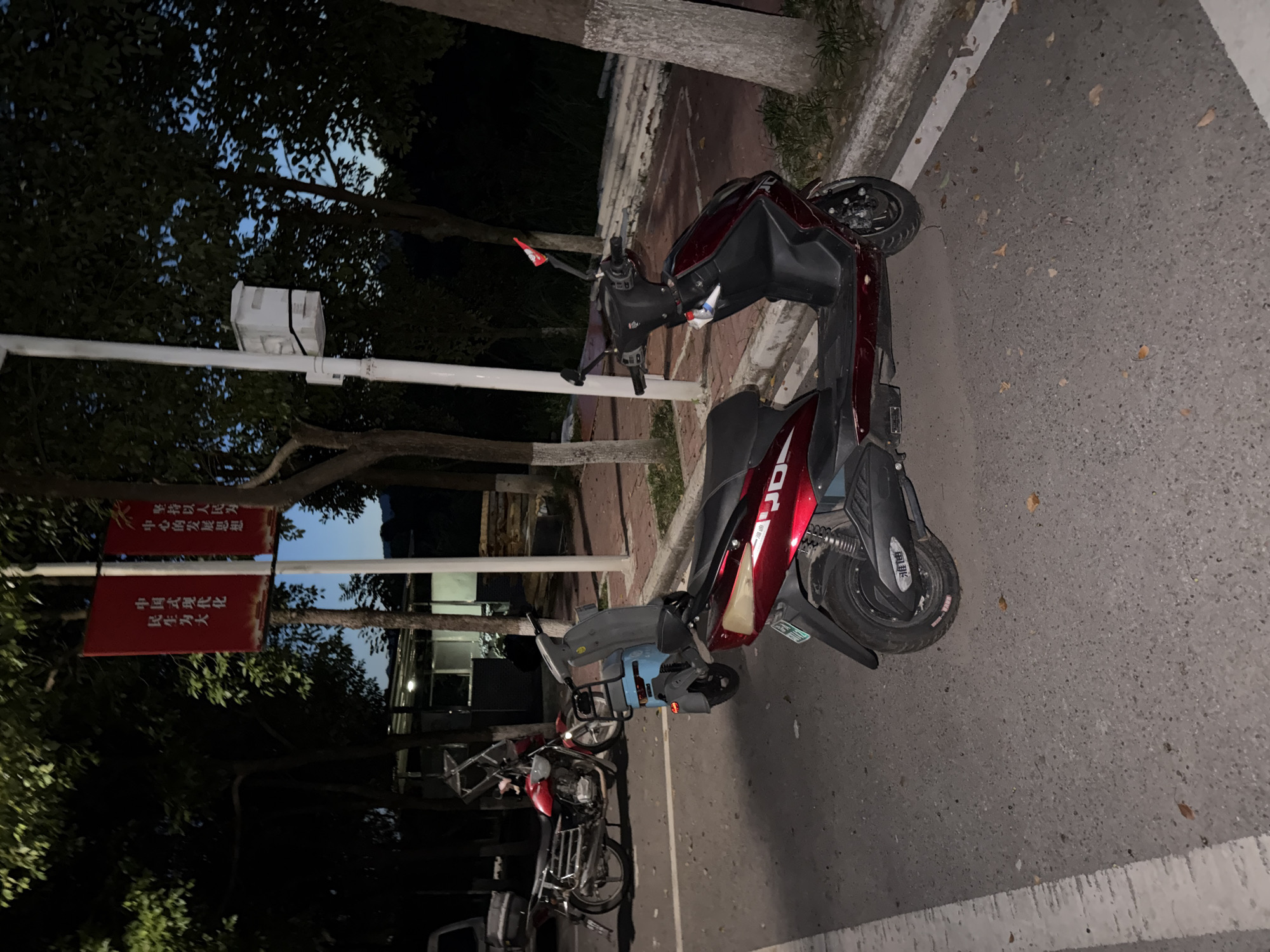 The image depicts an outdoor scene at dusk or night, likely in a public area of Jiangjin, China. In the foreground, a dark red electric scooter is parked on an asphalt road with white markings. The scooter has a black seat, black handlebars, and a black wheel well with white lettering. Behind it, a blue electric scooter is visible. Further back, a red and chrome motorcycle is parked. A tall white pole with a white box attached to it stands to the right of the scooters. Two red banners with Chinese characters are hanging from a pole to the left of the scooters, illuminated by an unseen light source. Trees with dark green foliage line the background, and the sky is a deep blue, indicating the transition from day to night. The ground is a mix of asphalt and paved areas, with some fallen leaves scattered about. The overall impression is one of a quiet street or public space after dark.