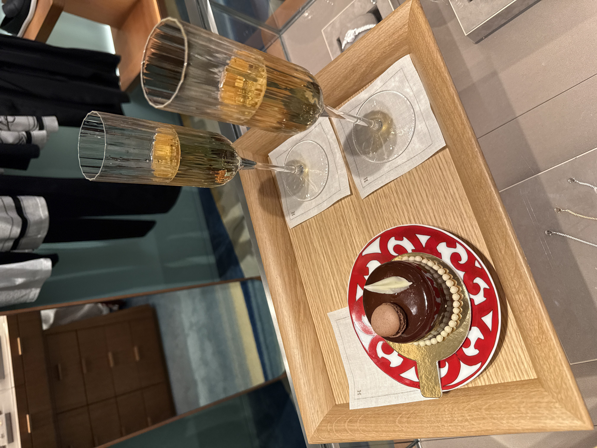 The image captures a luxurious scene set on a wooden tray, likely in a high-end boutique in Zapopan, Mexico. Two flutes filled with sparkling, light-golden liquid are prominently displayed, positioned towards the top and center-left of the frame. Below the flutes, on the right side of the tray, rests a small, elegantly presented chocolate cake on a decorative red and white plate. The cake is dark brown, glossy, and adorned with a white chocolate curl and a small, light brown macaron. White chocolate pearls encircle the base of the cake. Both the flutes and the cake are placed on small, white fabric napkins, each subtly embroidered with a distinct capital "H" logo, indicating a specific brand.
The wooden tray itself is light-colored with visible grain. In the background, reflections in what appears to be a mirror show parts of a room, possibly a changing area with folded clothing, and a wooden cabinet or shelving unit. To the right of the tray, a display area with a muted grey surface shows what looks like a silver necklace, suggesting a jewelry section. The overall arrangement suggests a moment of indulgence or hospitality offered to a customer, possibly during a luxury shopping experience. No people are visible in the frame. The lighting is soft and even, highlighting the details of the items.