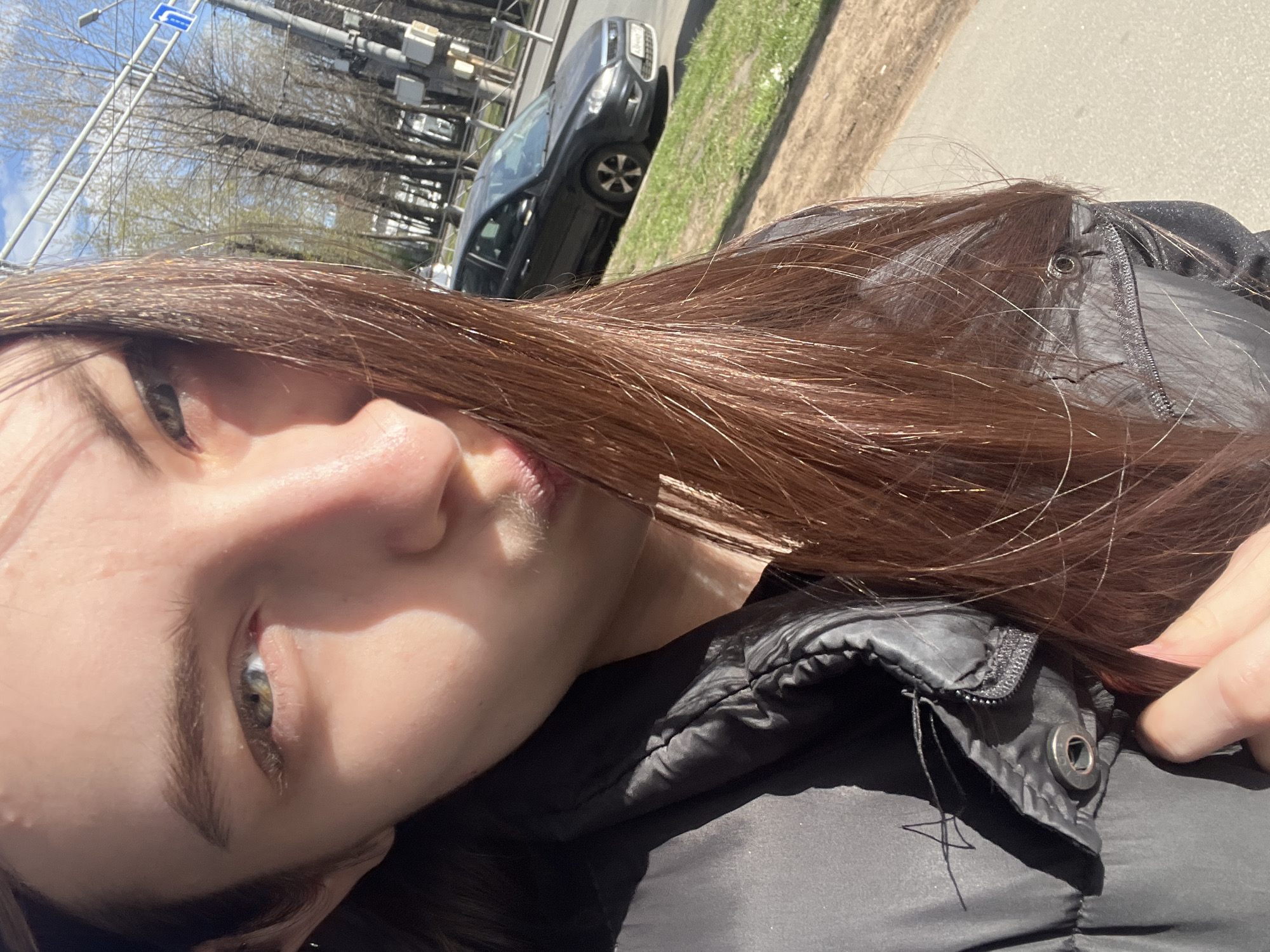 The image is an angled selfie of a young woman taken outdoors in what appears to be an urban or suburban environment in Saint Petersburg, Russia. The woman's face is prominent on the left side of the frame, with her left eye, nose, and part of her mouth visible as she looks towards the camera with a neutral expression. Her long, dark brown hair, illuminated by sunlight, cascades across her face and over her left shoulder. She is wearing a dark, possibly black, puffer jacket, detailed with a visible zipper and a metallic snap button near the collar. A hand is partially seen near her right shoulder, possibly holding her hair or adjusting her jacket.
The background, appearing rotated due to the selfie angle, shows a street scene under bright sunlight. A paved road is visible on the right, with a dark-colored car, likely a crossover or SUV, on it. Adjacent to the road are strips of green grass and a concrete or paved sidewalk/path. The sky is clear and bright blue with some white clouds. Numerous bare trees with visible branches suggest the season is either early spring before leaves have emerged or late autumn after they have fallen. Overhead infrastructure includes a utility pole or gantry supporting what appear to be street lights or traffic signals. A blue road sign with a white arrow pointing straight up, indicating "Go Straight" or "Ahead Only," is clearly visible.
The bright conditions indicate daytime. The scene captures a casual moment in a sunny, outdoor setting.