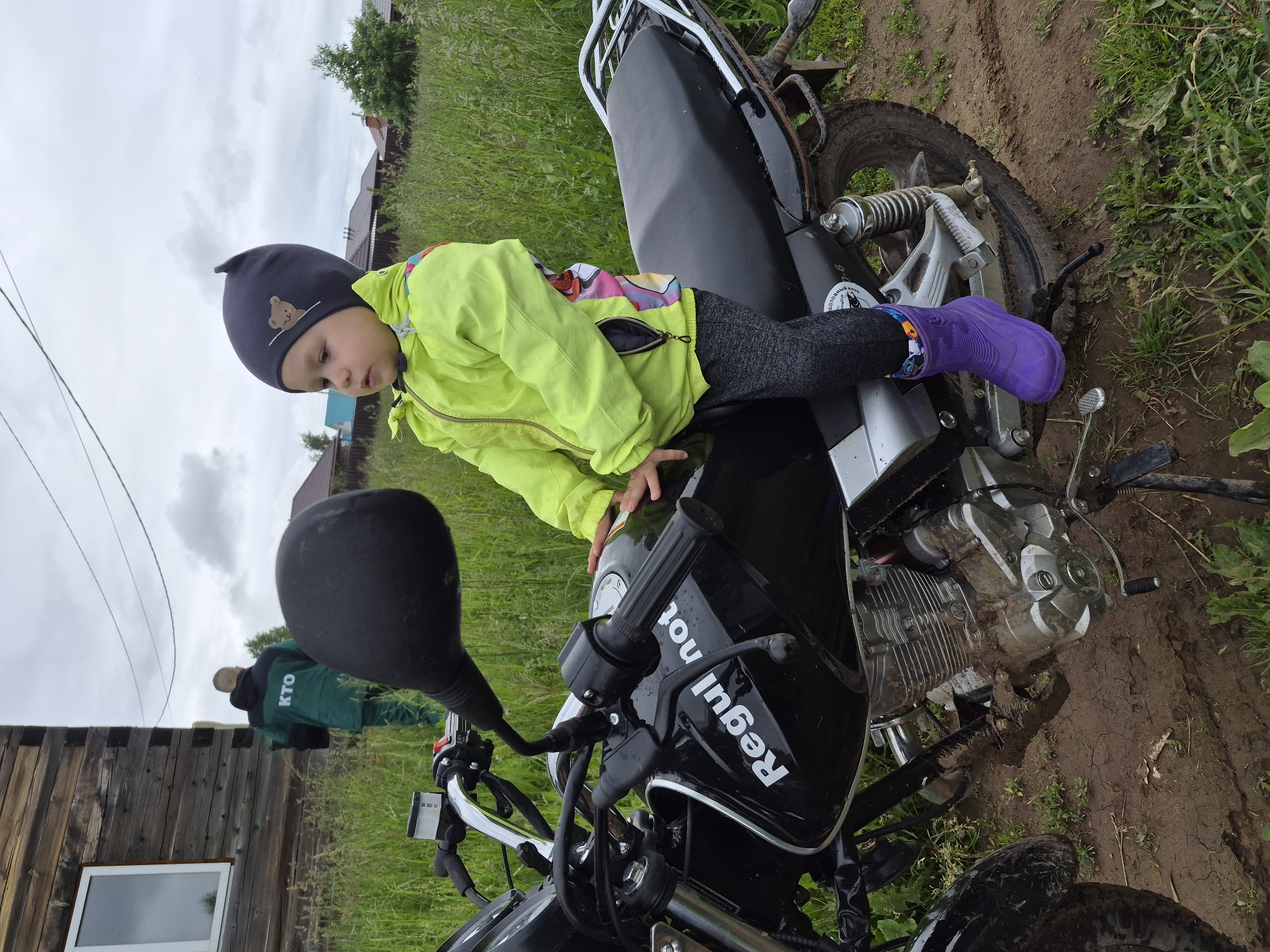 An outdoor scene in the city of Unknown, Unknown, depicts a young child seated on a black motorcycle on a muddy dirt path, with a rural backdrop. The child, facing slightly left, wears a dark grey beanie with a brown bear emblem, a neon yellow hooded jacket with colorful side panels, dark grey pants, and bright purple rubber boots. Their hands are positioned on the motorcycle's handlebars. The motorcycle, a black model with "Regul not" visible on the fuel tank, is extensively covered in mud, particularly around the engine and wheels. In the background, to the left, a person in a green jacket with "KTO" written in white on the back and matching green pants is walking away from a wooden log-cabin style building featuring a white-framed window. To the right, distant houses with dark roofs are visible on the horizon, interspersed with tall green grass and a small tree. Two thin utility lines traverse the overcast sky. The ground is characterized by churned mud and overgrown green grass.