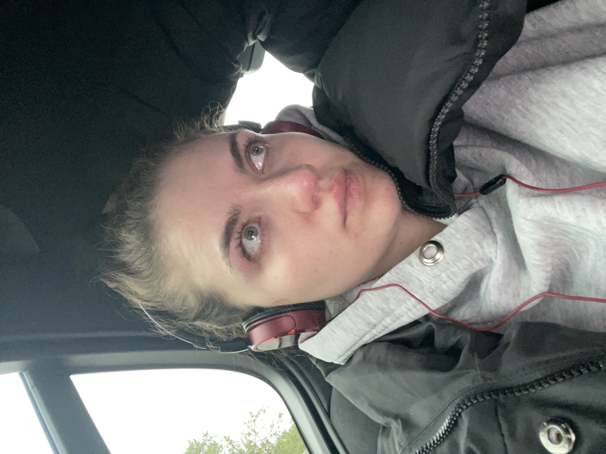 A woman is inside a car, with her face visible to the camera. She is wearing headphones, a grey hooded sweatshirt with red drawstrings, and a black puffer jacket. The interior of the car is visible, and outside the car, green foliage can be seen. The location is indeterminable without further context.