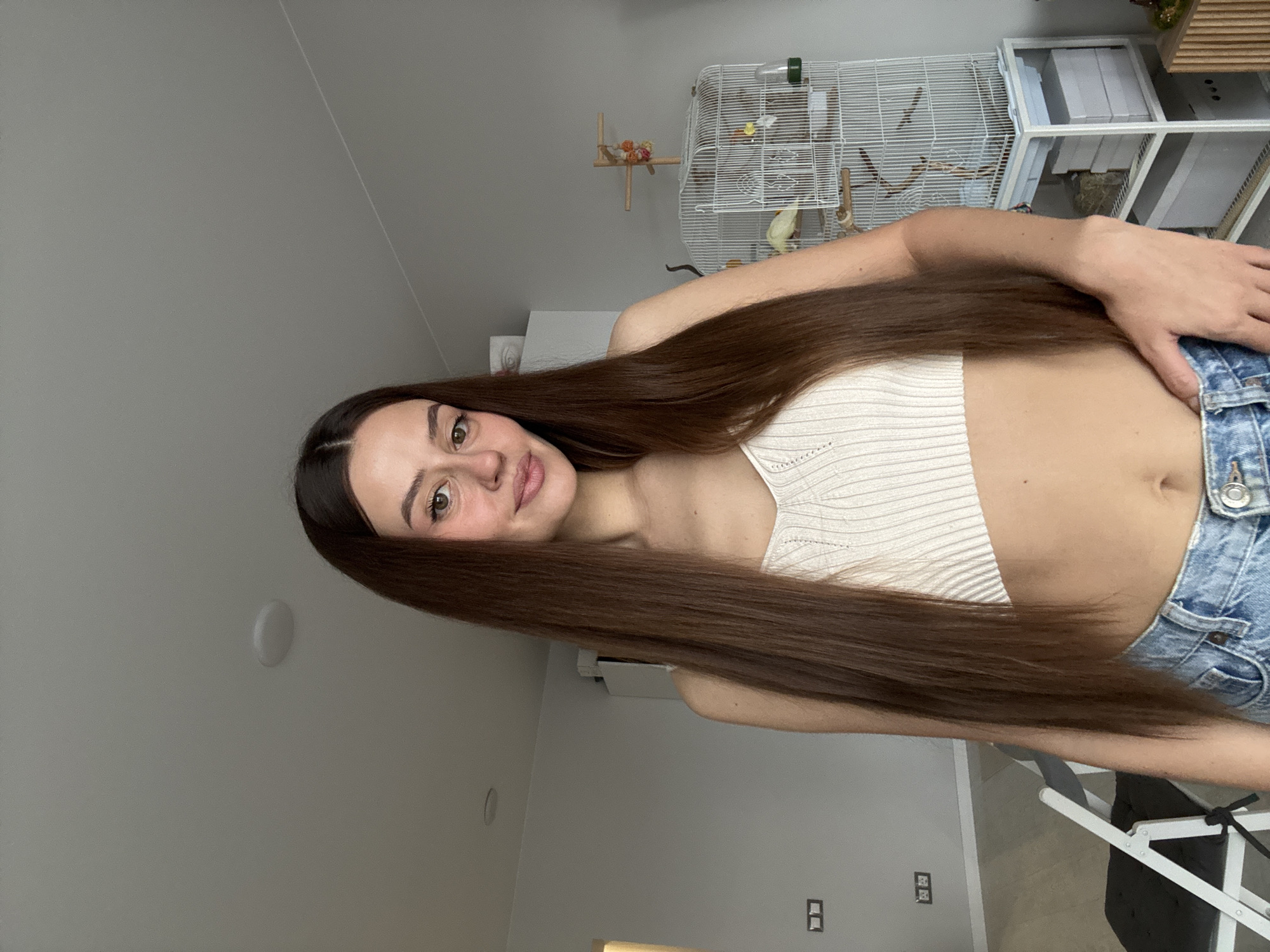A young woman with long brown hair is posing indoors. She is wearing a white crop top and denim shorts. In the background, a white birdcage with a bird inside is visible, along with a white shelf unit. The room is painted in light neutral tones. The location is likely a domestic residence, with light switches and electrical outlets visible on the walls. There is no definitive evidence within the image to pinpoint the location to Krasnoyarsk, Russia.