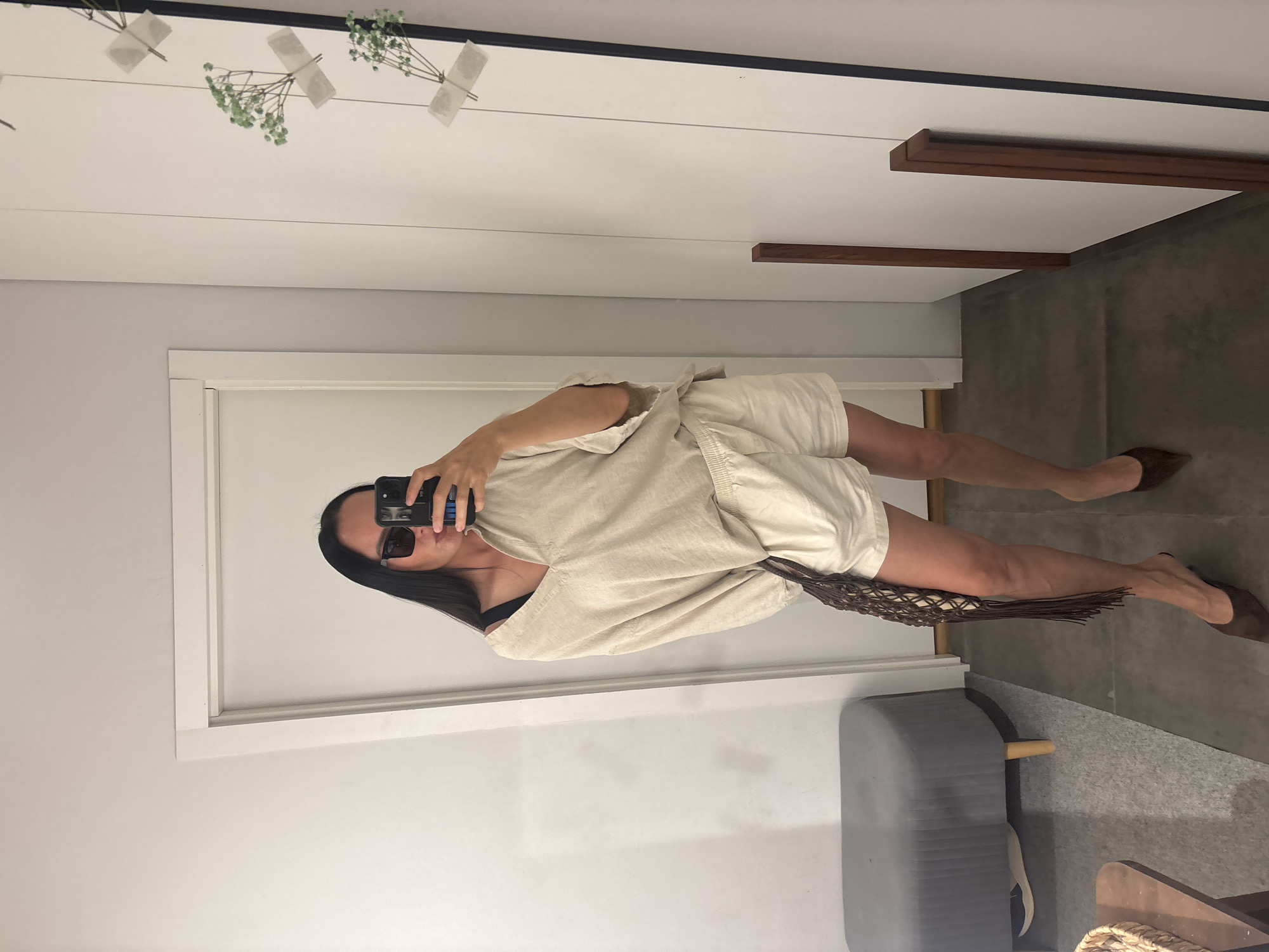 A woman is taking a selfie in a mirror. She is wearing a light-colored linen outfit, sunglasses, and brown shoes. She is also holding a brown net bag. The background includes a white wall, a doorway, a gray ottoman, and a glimpse of the floor. There are also some decorative plants taped to the wall. There is no discernible evidence in the image that indicates the location is the city of Chaikovsky, Russia.