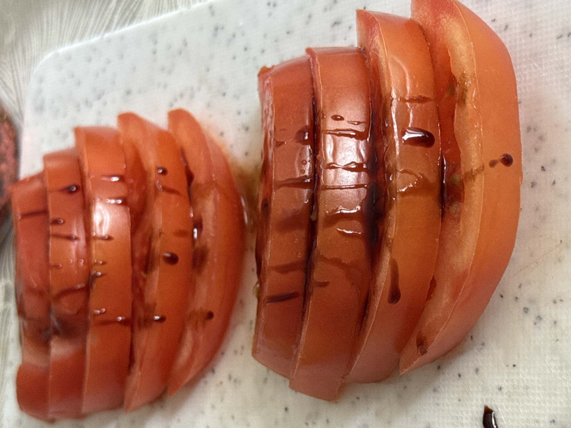 Two piles of sliced tomatoes arranged on a white speckled surface. Dark balsamic glaze is drizzled over the tomato slices. The scene's location is unknown.