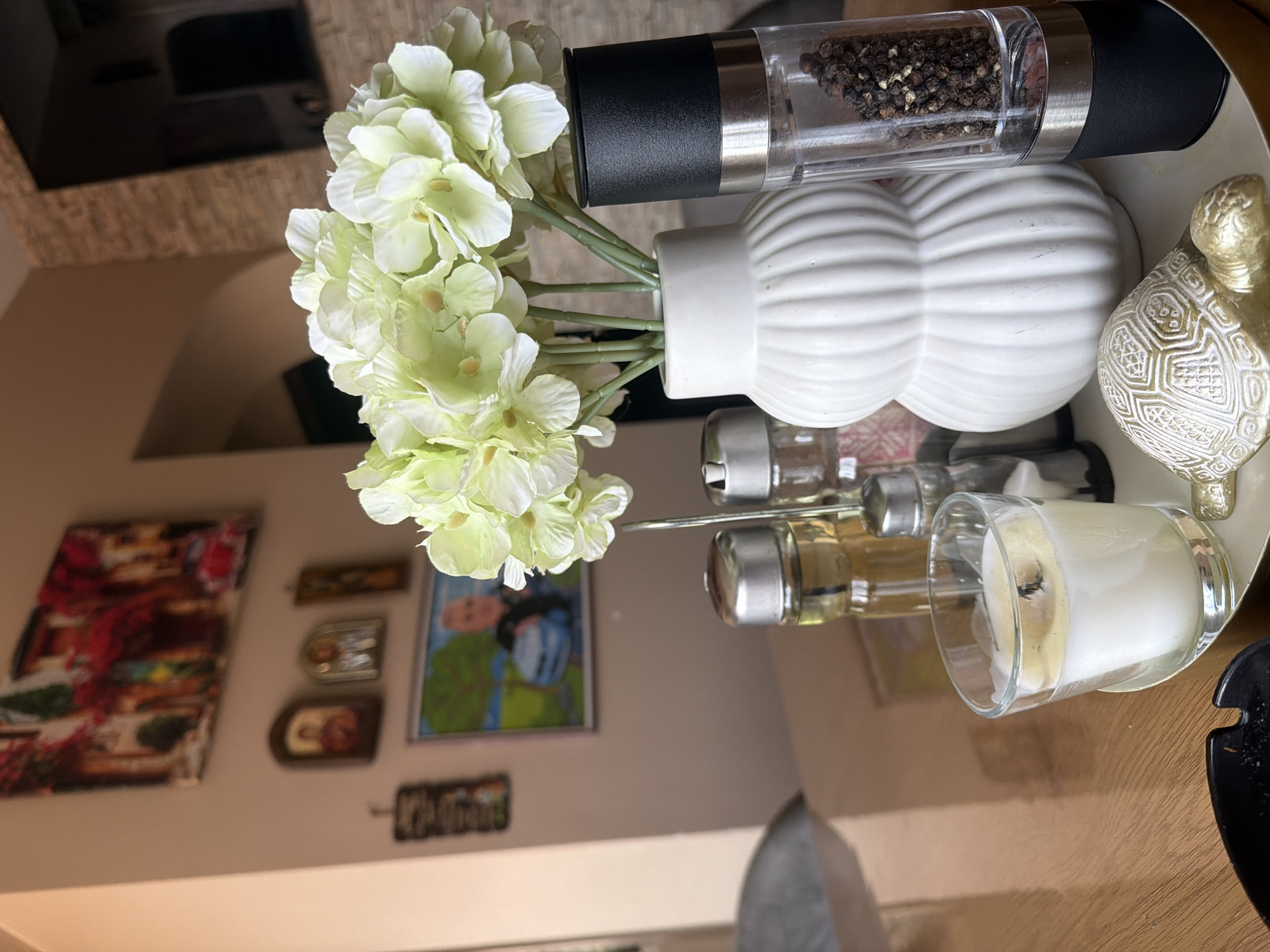 This indoor still-life photograph, taken in Shumen, Bulgaria, captures a decorative arrangement on a light-colored, possibly round or oval, tray. The image is taken from a slightly elevated, oblique angle, and appears to be rotated.
In the foreground, a vibrant bouquet of light green and white artificial hydrangea flowers stands in a small, white, fluted vase. To the right of the vase, a clear pepper grinder filled with black peppercorns features a black top and silver accents. Adjacent to these, a distinctive golden turtle figurine with intricate patterns on its shell adds an ornamental touch.
Further to the right and slightly below the pepper grinder, a clear glass votive candle holder contains a partially burnt white candle, indicating prior use. Next to the candle, several small glass bottles, possibly containing oils or spices, are visible on the tray.
The background reveals a room interior. On a light-colored wall, several framed pictures are displayed. One clearly visible framed image depicts a person, possibly a child or young adult, wearing a blue helmet and shirt, set against a green, possibly natural, background. Other smaller frames and what appears to be a piece of text art are also on the wall, though the text itself is largely illegible. Above the wall art, a wooden architectural element, possibly a ceiling beam or part of a furniture piece, is visible. The overall setting suggests a cozy and decorated living space. No specific activity is occurring, presenting a static snapshot of an organized display.