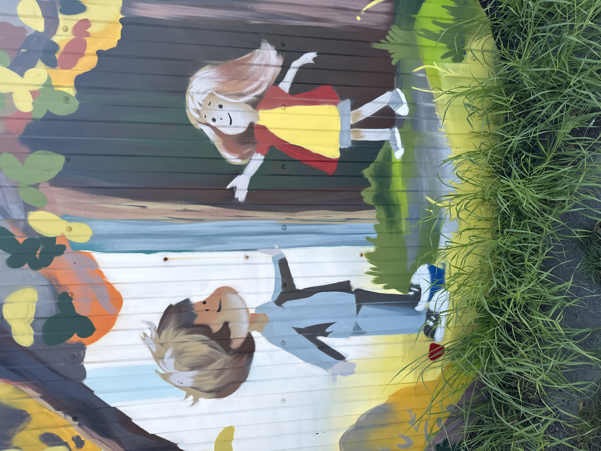 The image depicts a colorful mural painted on a corrugated metal surface, likely an outdoor fence or wall. The mural features two cartoon-style children standing in a stylized forest setting. On the left, a boy with light brown hair and a blue outfit is depicted. His arms are outstretched to the sides, and his hands are open. To his right, a girl with light brown hair, a yellow dress, and a red jacket is shown with her arms extended outwards. Both children are facing each other. The background includes abstract representations of trees, leaves, and grass, with a bright yellow and green color palette dominating the lower portion. In the foreground, tall green grass partially obscures the bottom of the mural and the ground. The mural is located in the city of Rusanovo, Russia.