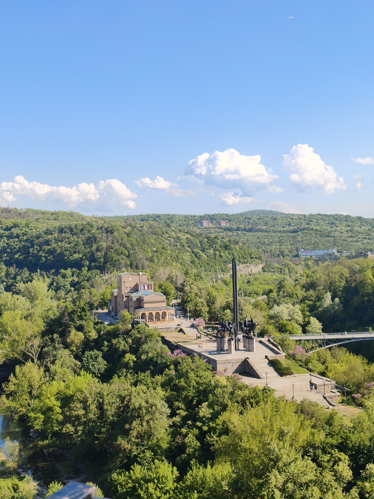 An eye-level, wide, outdoor shot depicts a monument and museum atop a plateau surrounded by lush, green vegetation in Veliko Tarnovo, Bulgaria.
In the midground, the Mother Bulgaria Monument stands tall with a central dark-colored obelisk surrounded by figurative sculptures. Adjacent to the monument, a tan-colored building with arched openings is partially visible. A light-colored bridge spans the landscape, connecting the plateau to the right side of the frame.
The foreground is filled with vibrant green trees and foliage, partially obscuring the view of the river below. The background displays a dense forest stretching towards the horizon under a blue sky dotted with fluffy white clouds. The overall environment suggests a serene, natural setting integrated with historical and cultural landmarks.