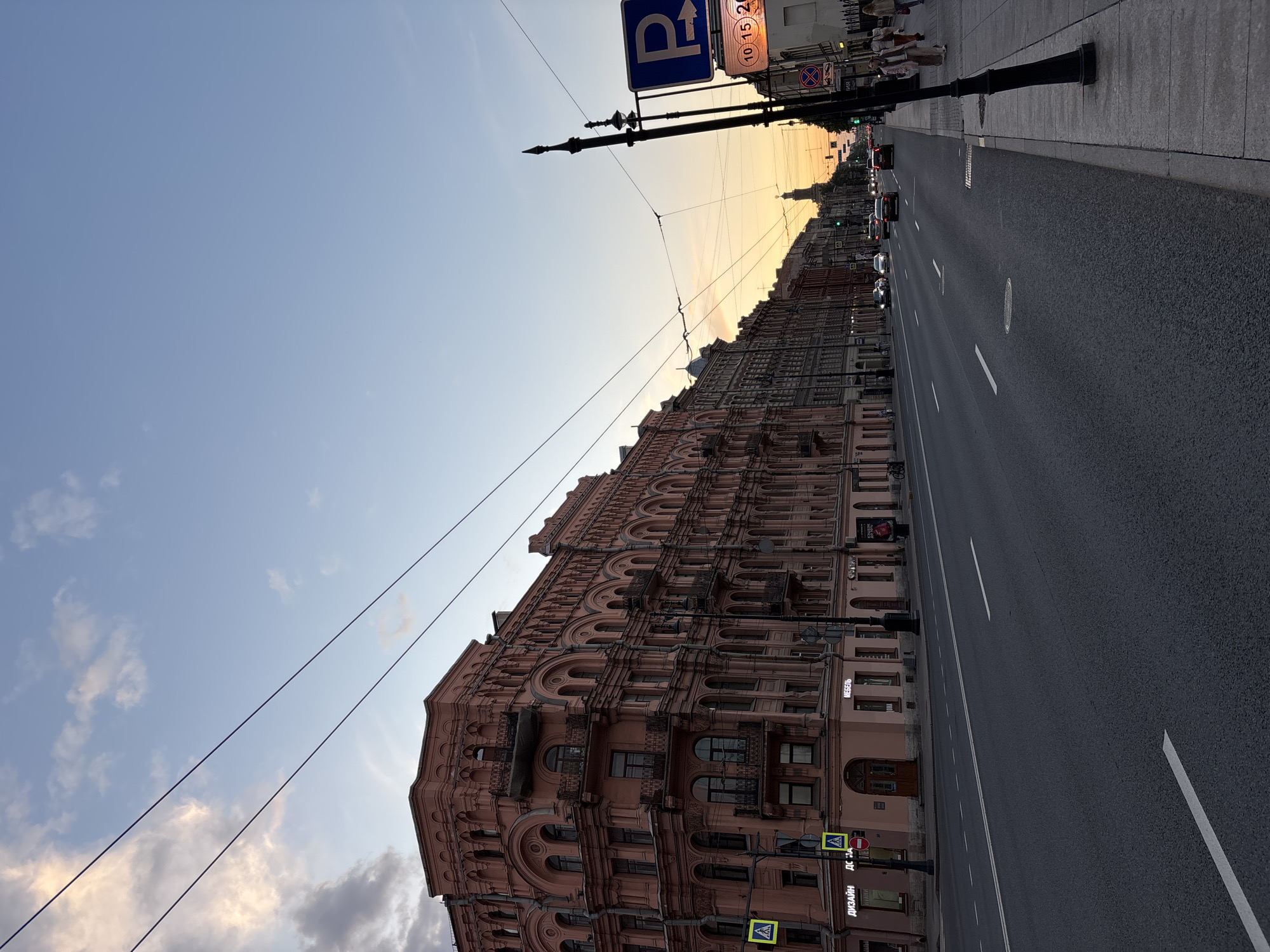 The image captures a wide city street in Saint Petersburg, Russia, at what appears to be either sunset or sunrise, given the warm orange glow along the horizon line transitioning to a pale blue sky with scattered clouds overhead.
On the left side of the street, a long, ornate multi-story building with a pinkish or reddish facade dominates the view. It features numerous arched windows and decorative balconies on its upper floors. At street level, shops are visible, including one with Cyrillic text "МЕБЕЛЬ" (FURNITURE) and another stating "ДИЗАЙН ДС" (DESIGN DS). Yellow pedestrian crossing signs are also affixed to the building's base.
The paved road occupies the center and right side of the frame, showing white lane markings. Several cars are visible in the distance, heading away from the viewer. On the right-hand sidewalk, a classic black lamppost stands, and a few people can be seen walking further down the street. Overhead, power lines or trolleybus wires stretch across the scene.
Prominently displayed above the right sidewalk is a blue rectangular parking sign with a white 'P' and an arrow pointing right. Below the 'P', smaller text "10-15-20" is visible, likely indicating parking hours or rates. Another circular red sign with a diagonal line (typically signifying 'No Stopping' or 'No Parking') is also present. The overall lighting is dim, emphasizing the dramatic sky.