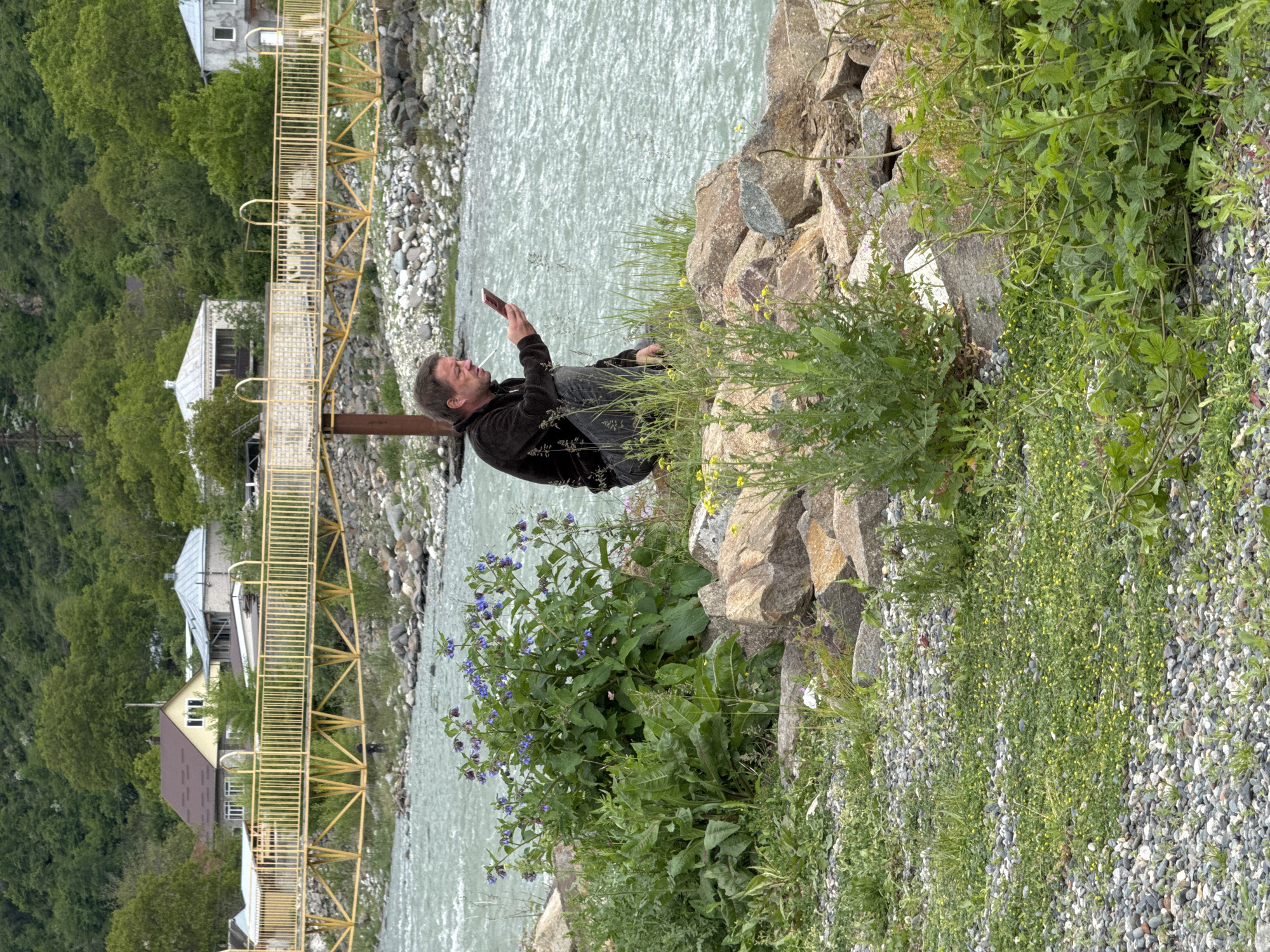A man reclines on rocks beside a turbulent river in Karachayevsk, Russia, engrossed in his mobile phone. He wears a dark jacket and gray trousers, his posture relaxed as he interacts with the device. The river, characterized by its grayish-white color and rapid flow, contrasts with the adjacent yellow pedestrian bridge, which leads to houses nestled among lush green trees. The foreground consists of a mix of rocks, grass, and blue flowers. The overall scene presents a moment of technological immersion amidst a natural, rugged landscape.