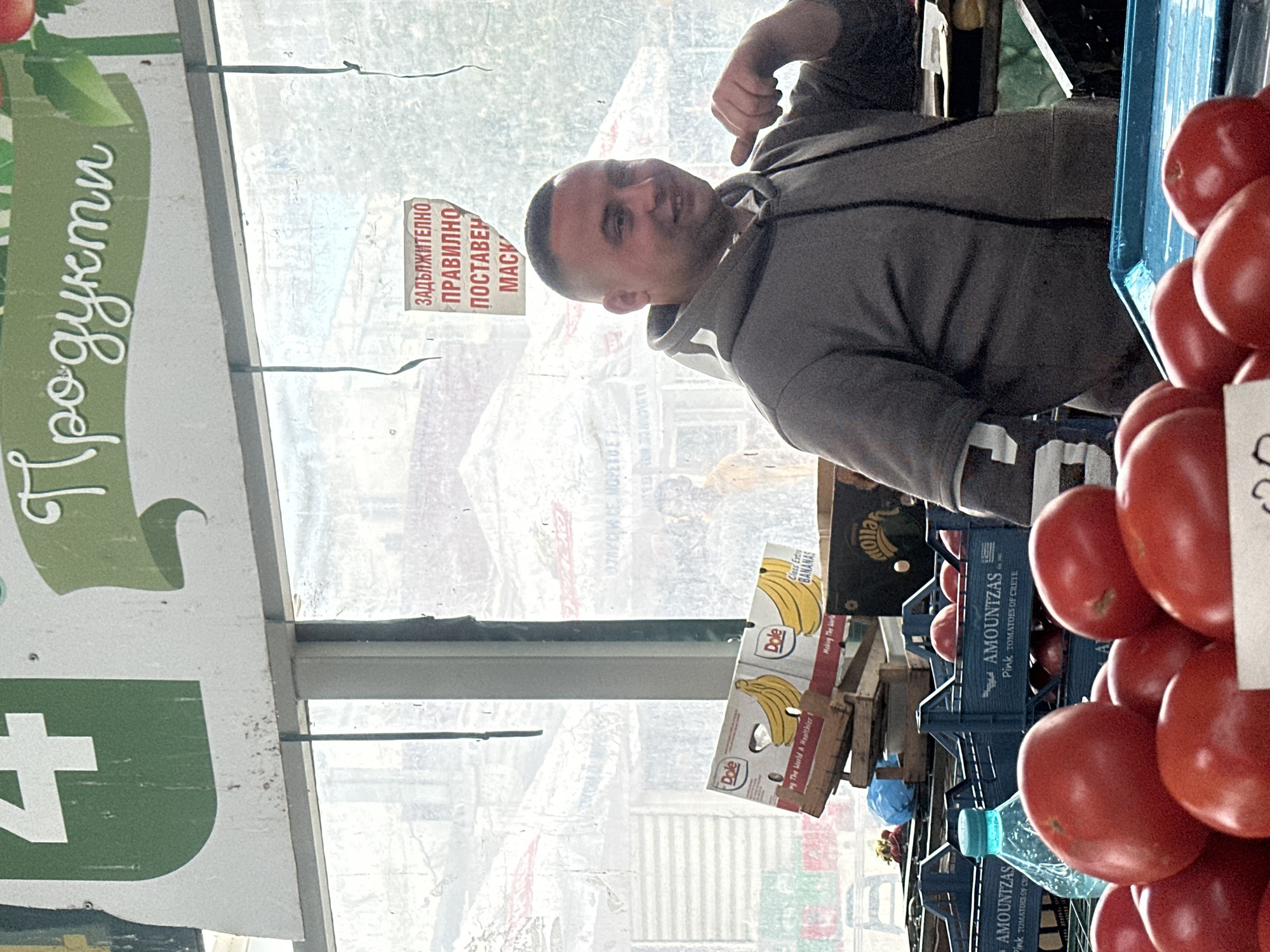 The image shows a man standing at a fruit and vegetable stand, likely at a market, with the word "Продукти" (products) visible in Bulgarian on a sign. There's another sign that says "ЗАДЪЛЖИТЕЛНО ПРАВИЛНО ПОСТАВЕН MACK" (Obligatory to wear a mask properly). Boxes of Dole bananas and blue crates of "AMOUNTZAS Pink TOMATOES OF CRETE" are visible. The background shows an urban environment seen through a window, potentially locating the image in Sliven, Bulgaria, based on the Bulgarian language signs. The man is wearing a gray hooded sweatshirt and appears to be gesturing towards the viewer. Stacks of tomatoes are visible in the foreground.