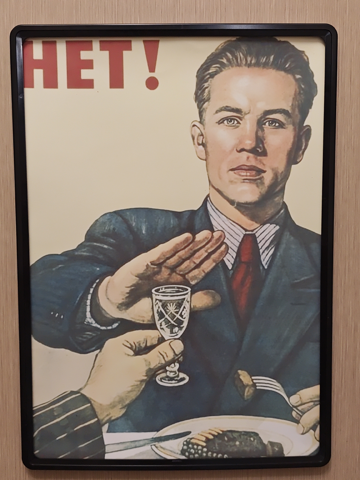A framed Soviet-era anti-alcohol poster depicts a man in a dark suit and red tie pushing away a shot glass of alcohol offered by another person's hand. The man's expression is firm, and his hand is held up in a gesture of refusal. In the foreground, a plate of food, possibly meat and berries, is visible, with a fork spearing a piece of meat. The background is a plain, light color. The poster reads "HET!" in large red letters at the top, which translates to "NO!" in Russian. There is no discernible location in Saint Petersburg, Russia, indicated in the image.