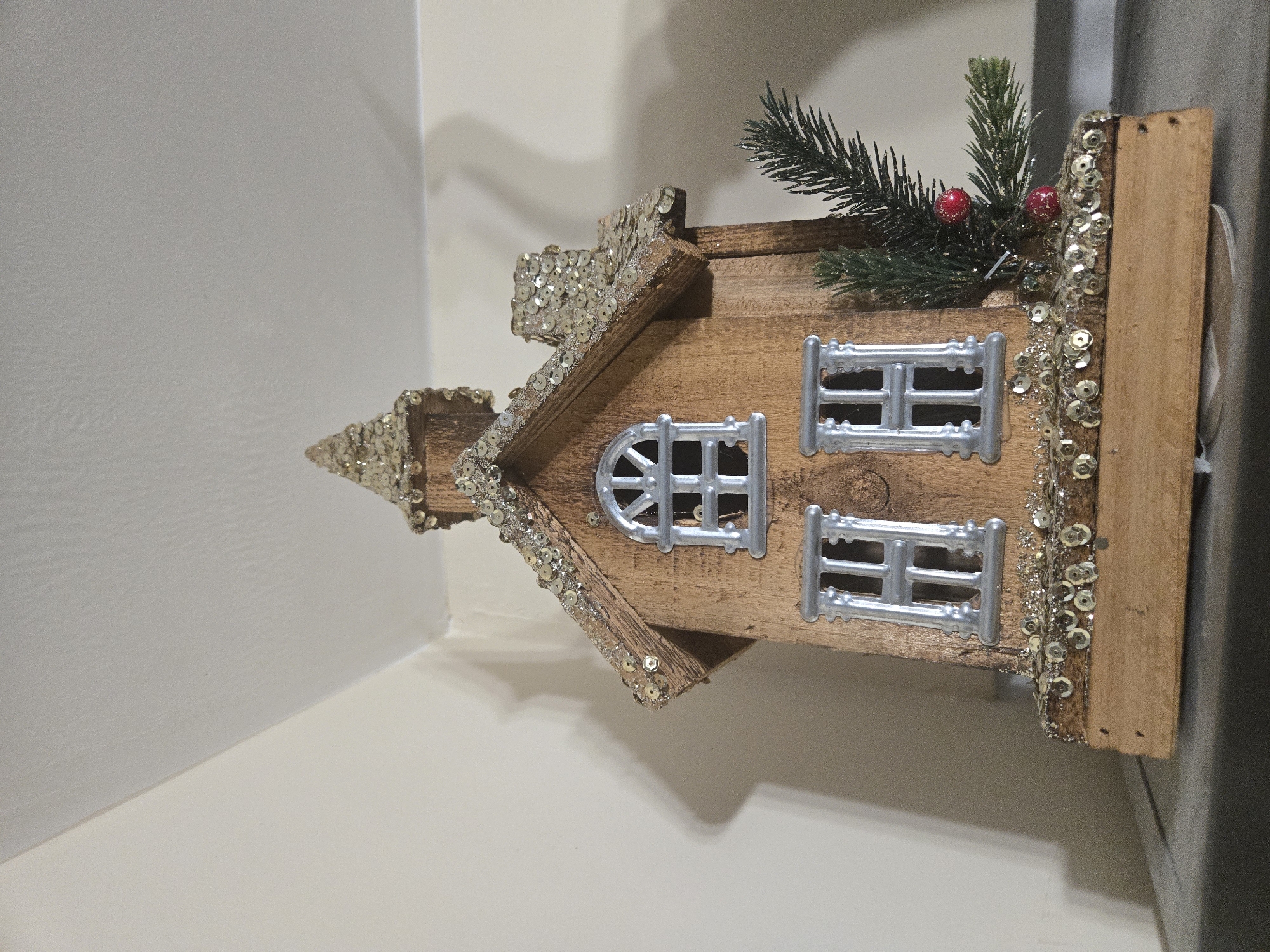 A decorated wooden house miniature is positioned in a corner where two walls meet. The miniature house is made of light brown wood with silver colored plastic windows and a coating of silver sequins along the roof edges, chimney, and side wall. It also has a small artificial pine sprig with red berries on top of the side wall. The walls are off-white. The location is unknown.