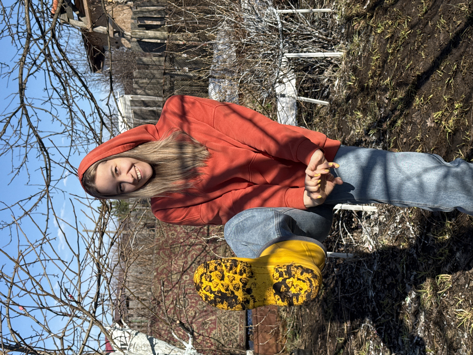 A young woman in an orange hoodie and blue jeans stands outdoors in Chaikovsky, Russia. She wears yellow rain boots caked with mud. The background includes bare trees and a glimpse of buildings, with the ground covered in patchy grass and soil.