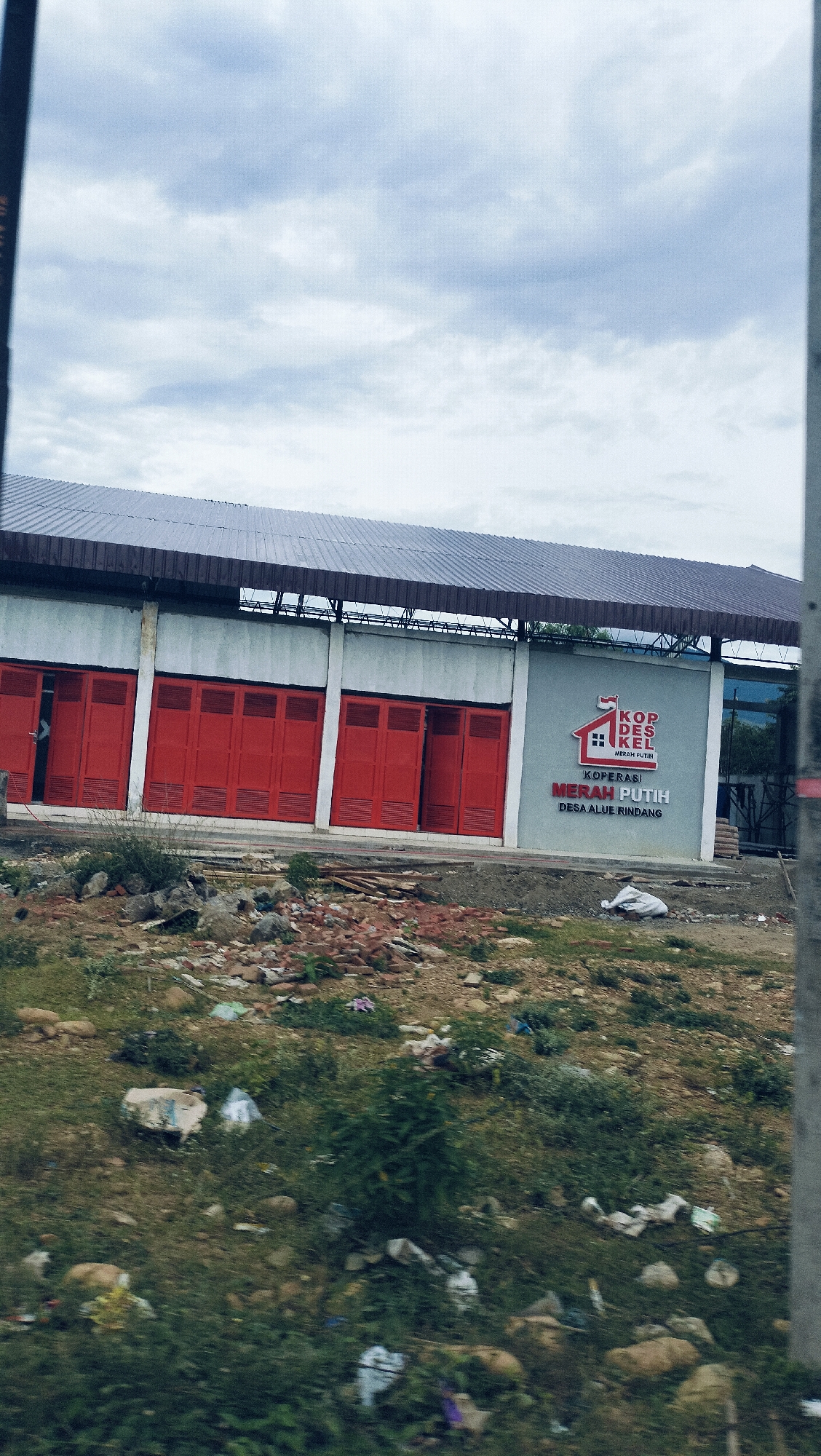 The image displays an exterior view of a building under an overcast sky, likely in Lonbaroh, Indonesia. The building has light grey walls and a dark, possibly corrugated metal roof. The left side of the facade features a series of five prominent red folding doors or shutters. To the right of these doors, a section of the wall serves as a large sign.
The sign prominently features a logo depicting a stylized house with a red and white roof, resembling the Indonesian flag. Above the house, the text "KOP DES KEL" is visible, with "MERAH PUTIH" in smaller red letters underneath. Below the logo, larger red text reads "KOPERASI MERAH PUTIH," and beneath that, in smaller grey letters, "DESA ALUE RINDANG." "Koperasi Merah Putih Desa Alue Rindang" translates to "Red White Cooperative of Alue Rindang Village."
The foreground and midground in front of the building consist of an uneven, unpaved area. This ground is sparsely covered with grass and dirt, and is littered with various debris including rocks, broken bricks, pieces of wood, and scattered plastic waste. This suggests a raw, undeveloped, or possibly neglected area. No individuals or specific activities are visible. The sky is uniformly covered with light grey clouds, indicating an overcast or cloudy day. The overall scene portrays a cooperative building in a somewhat rural or developing setting.