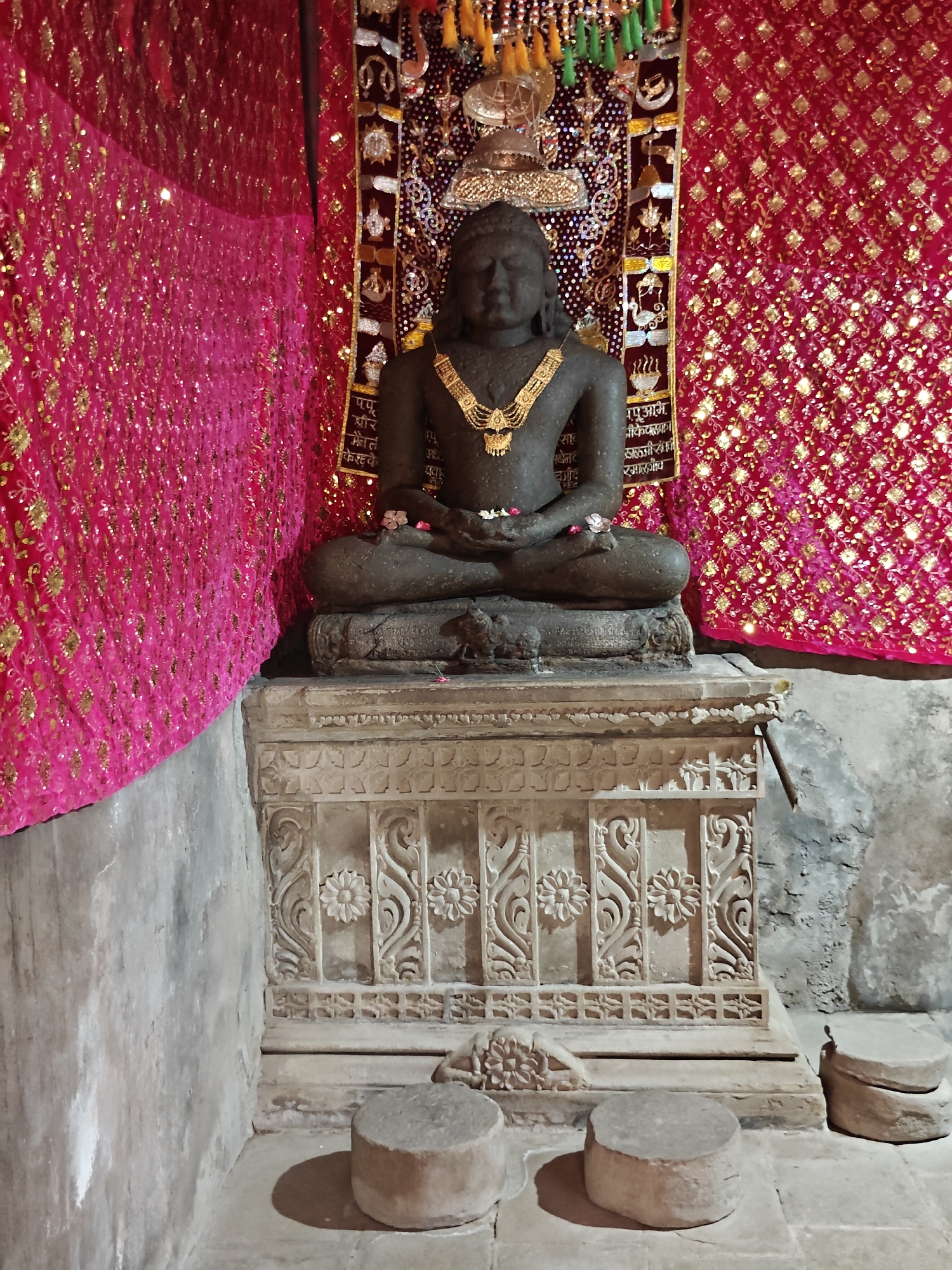 The image presents a statue in a room, potentially a shrine or temple setting. The statue is a dark stone figure seated in a lotus position on a decorated pedestal. It is adorned with a gold necklace. There are two small flowers near the statue's hands. Behind the statue is a red and gold decorated backdrop. To the left, the wall is covered with pink fabric. In front of the pedestal, there are two cylindrical stone objects that appear to be used as seats. The location is identified as Kaswara, India.