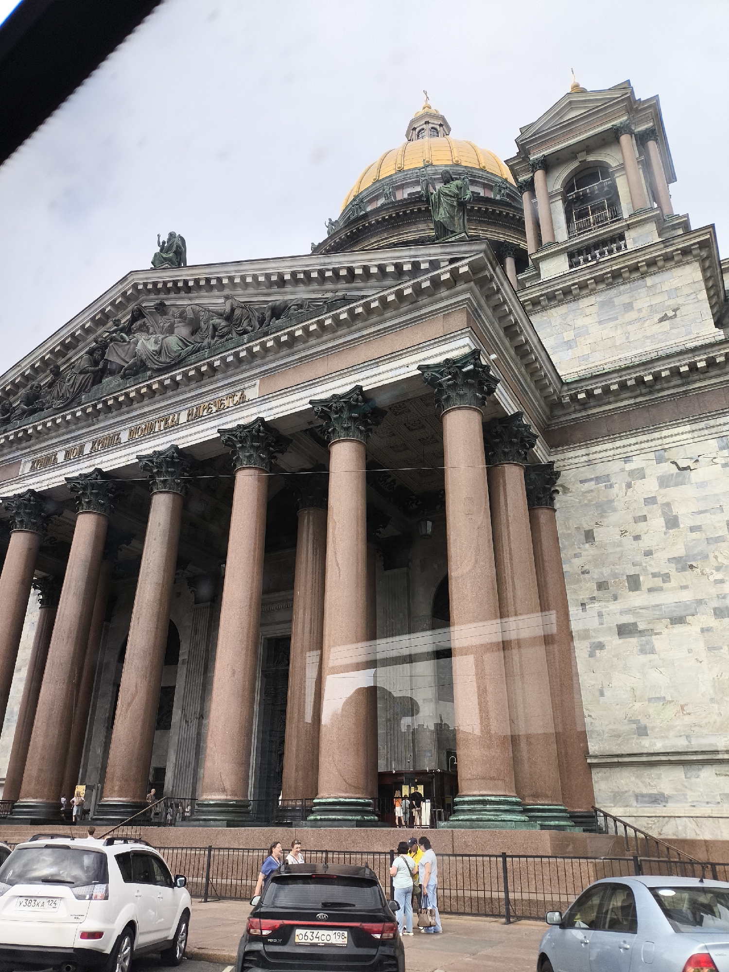 The image shows a frontal view of a grand cathedral with a golden dome, likely St. Isaac's Cathedral in Saint Petersburg, Russia. The cathedral features massive pink granite columns supporting a stone facade adorned with sculptures and an inscription. The sky is overcast, suggesting a cloudy day. In the foreground, several cars are parked, including a white SUV and a black Kia. A few people are visible near the cars and the entrance to the cathedral, some appearing to be tourists. The reflection of the sky and possibly other buildings is visible on the glass of the cathedral's entrance. The overall scene depicts a tourist visiting a historical landmark. The text on the facade of the cathedral is partially visible and appears to be in Cyrillic.