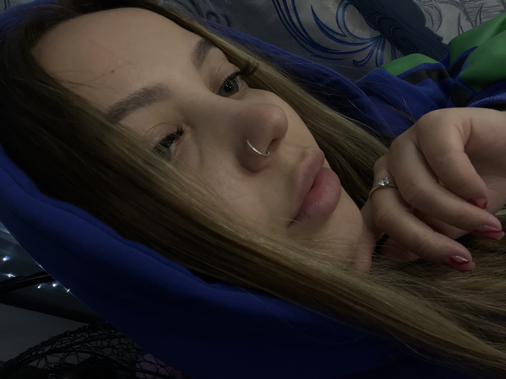 A close-up shot of a woman with light brown hair, wearing a blue hooded sweatshirt, resting her head. She has a nose ring and is wearing a ring on her finger. Her nails are manicured with a red design. The background is blurred, showing a patterned fabric and some lights. The location, Novopokrovskaya, Russia, is not discernible from the image.