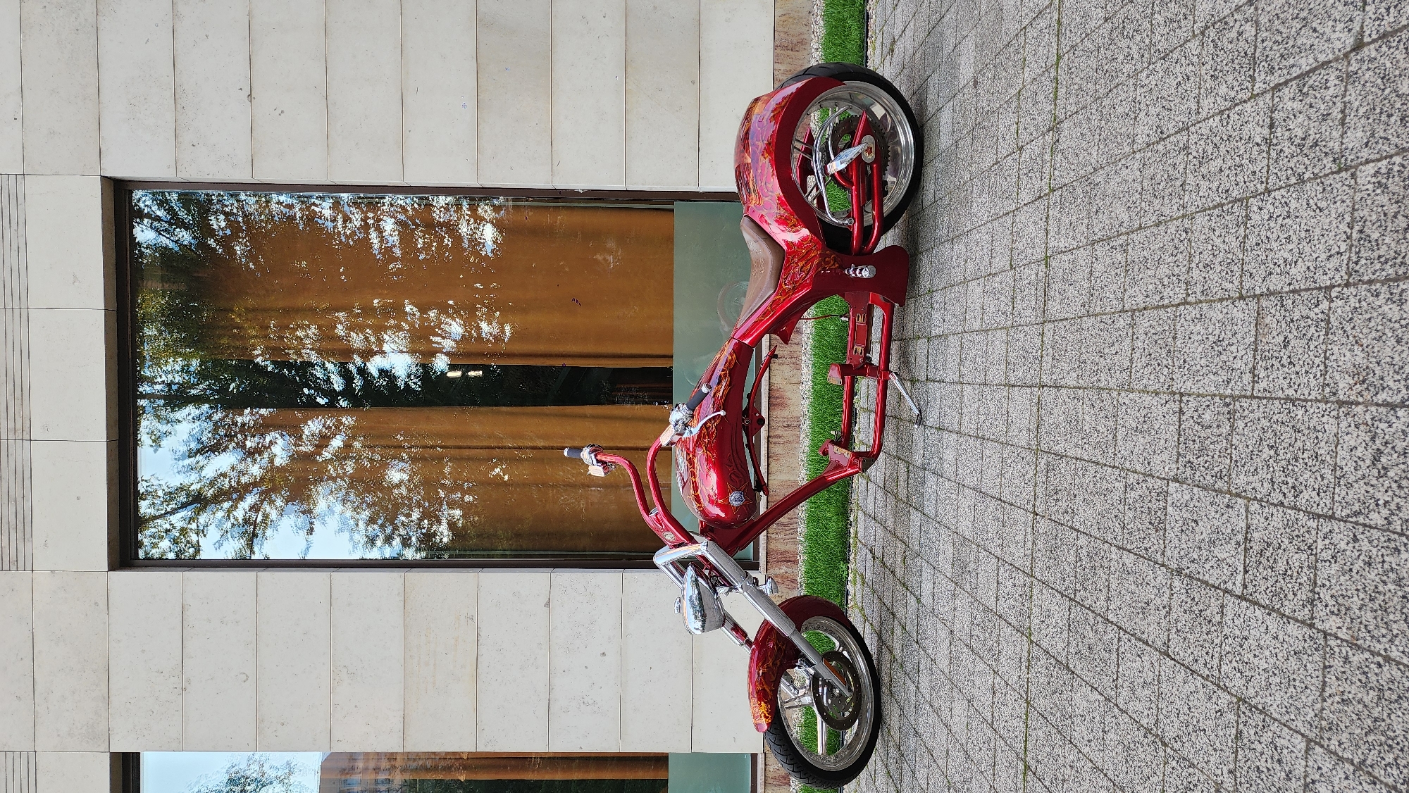 A custom red motorcycle is parked on a patterned stone sidewalk next to a building with large windows and a grassy strip. The motorcycle is mostly red with flame-like decals. It has a prominent front wheel with spokes and a chrome fender. The handlebars are visible, as is the seat and the frame. The motorcycle's rear wheel is also visible, with a large brake disc. The building has light-colored stone facade and large windows that reflect trees and the sky, suggesting it is daytime. The image is oriented vertically, with the motorcycle appearing to lean to the right. The sidewalk is made of grey square tiles with a speckled pattern. The overall scene is well-lit, implying a clear day. No people are visible. The location context is Moscow, Russia. There is no visible text in the image.