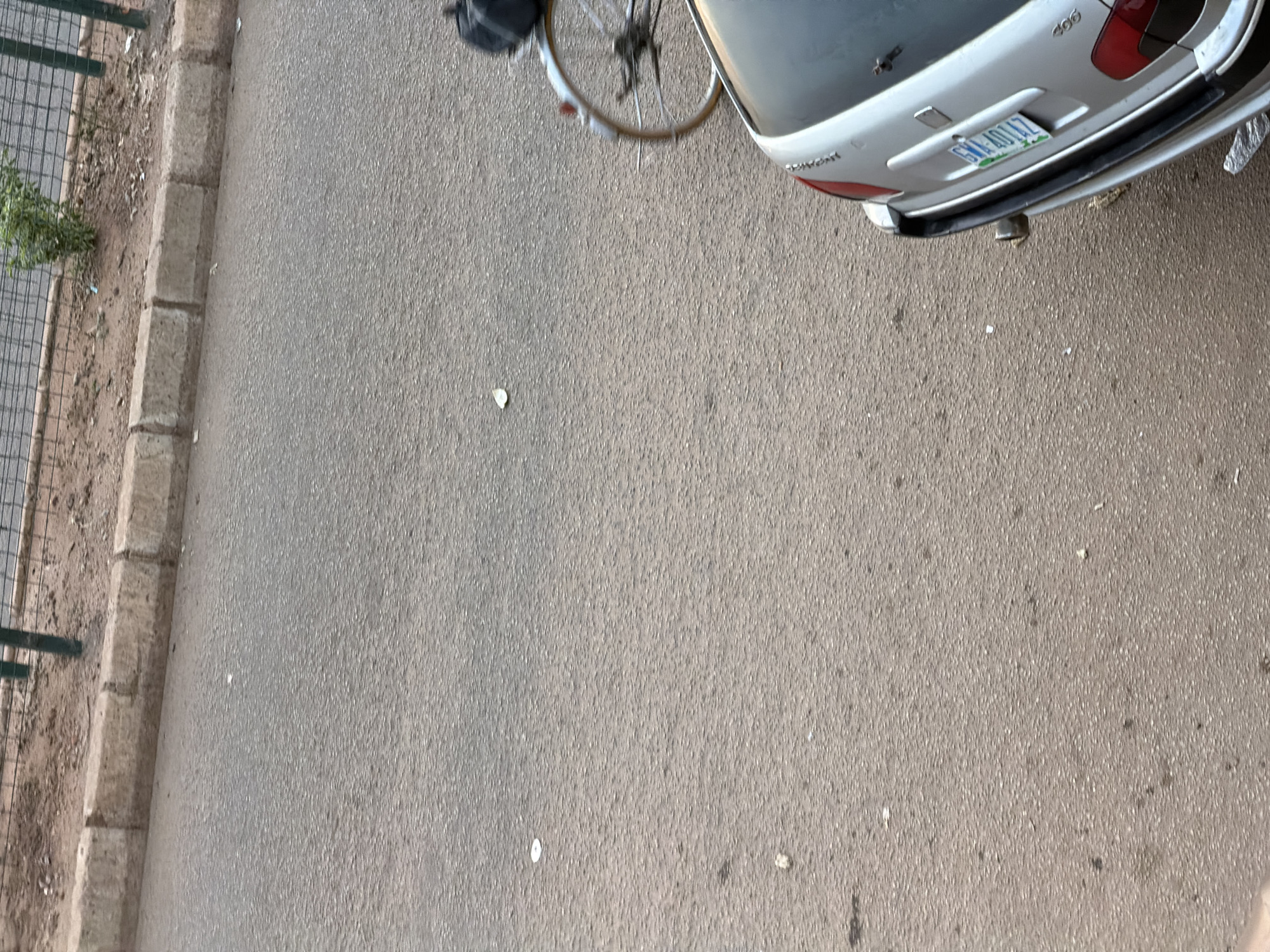 A low-angle, medium shot captures a segment of a paved road and sidewalk in Bukuru, Nigeria. On the right side of the frame, the rear of a silver sedan is visible, its license plate reading "GMA 4014Z". To the left of the car, a bicycle with a dark bag on its rear rack is partially visible. The road surface is asphalt, with scattered debris. A concrete curb separates the road from a dirt area with sparse vegetation and a metal fence at the top of the image.