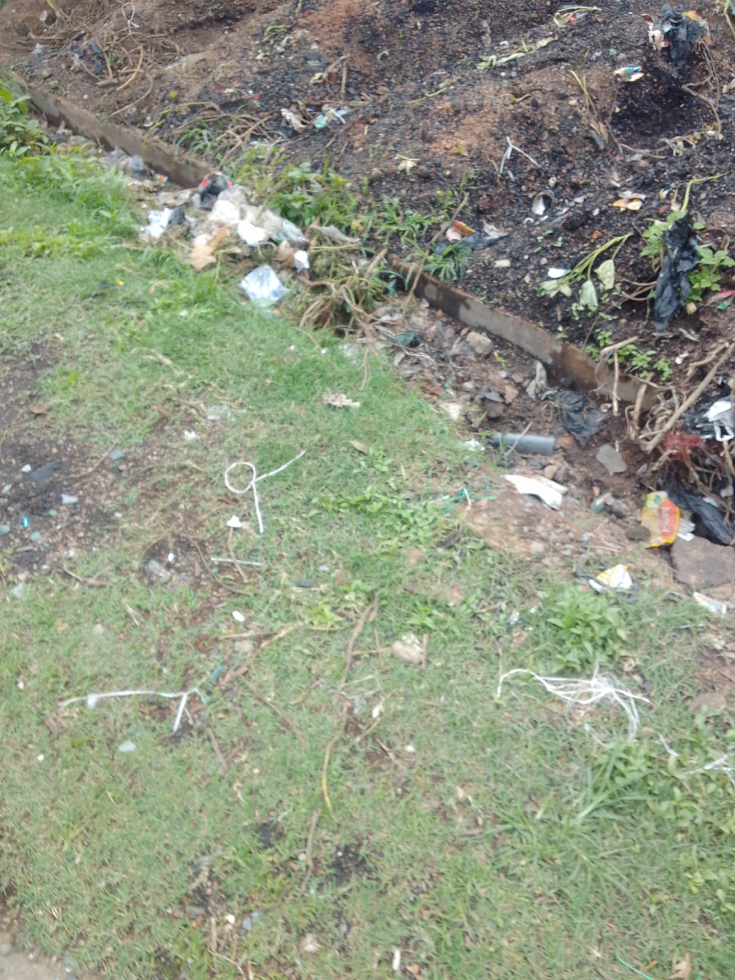 The image depicts a static, ground-level scene of a neglected outdoor area in Jos, Nigeria, characterized by a mixture of sparse vegetation, exposed soil, and significant refuse accumulation.
The scene is divided by a horizontal, linear barrier, likely concrete or wood, running across the middle. Above and to the left of this barrier, the ground is covered by patches of short green grass intermingled with bare earth. Several pieces of white string or thin rope are scattered across this grassy section, with one distinct loop formation visible in the central-left.
Below and to the right of the barrier, the terrain transitions into a lower-lying trench or ditch area composed primarily of dark, disturbed soil. The upper right portion of this dirt area shows evidence of charred material or ash. This trench is heavily inundated with various types of solid waste, including numerous crumpled white and black plastic bags, colorful plastic wrappers, fragments of paper or cardboard, and scattered organic debris. A segment of a grey cylindrical pipe, possibly PVC, is partially visible within this accumulated trash in the lower central-right. Dry branches, twigs, and sparse, struggling green weeds are also present, integrated with the soil and refuse.
No individuals, animals, or active movements are depicted in the scene. The lighting suggests daylight conditions with soft diffusion.