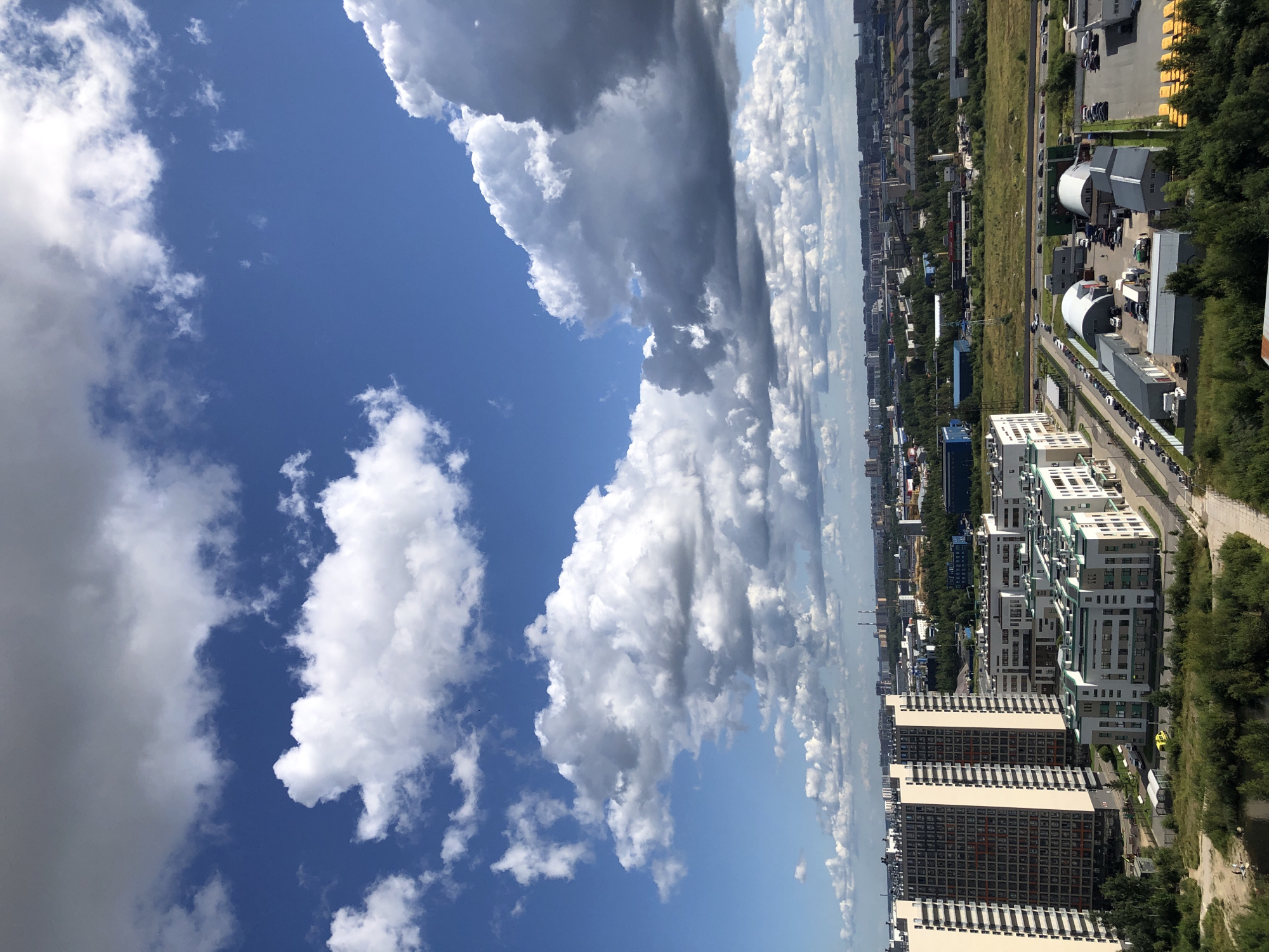 An aerial, full shot captures a cityscape under a bright blue sky dotted with voluminous white and gray clouds. In the foreground, a dense line of lush green trees partially obscures the lower edge of the frame. To the left, a tall, modern residential building with a dark, patterned facade and light-colored vertical accents dominates the view. Adjacent to it, a cluster of lower, contemporary buildings features white facades and green trim. Further into the mid-ground, a long stretch of road is visible, lined with parked cars and a row of yellow school buses. Several industrial-style buildings with rounded roofs and loading docks are also present. The background reveals a sprawling urban landscape with numerous buildings of varying heights, interspersed with patches of greenery and industrial structures. The overall impression is of a sunny day in an active, developing urban area.