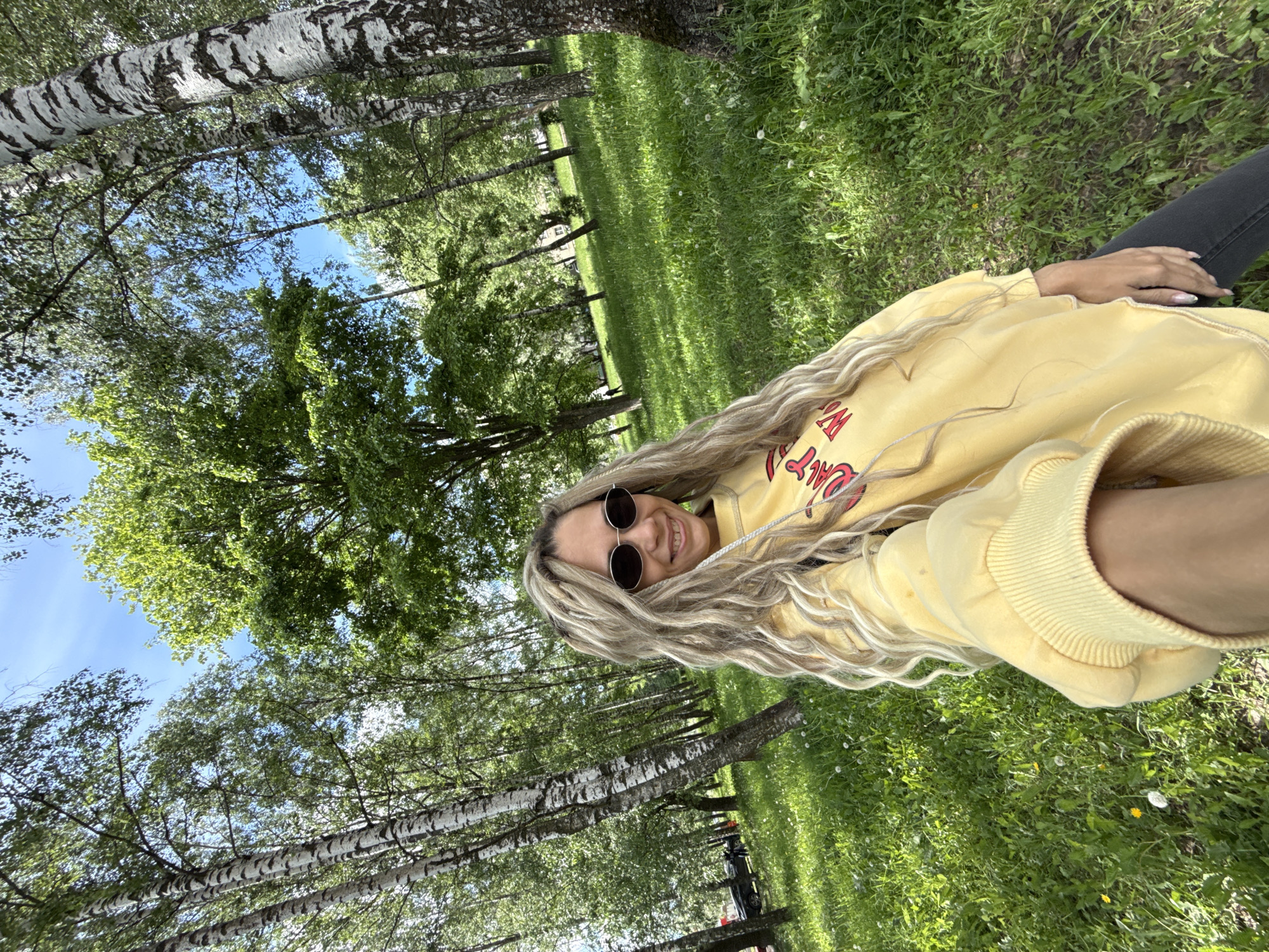The image shows a person lying on their side in a grassy area surrounded by birch trees. The person has long, blonde hair, is wearing sunglasses, and a yellow sweatshirt. The location appears to be a park or green space, possibly in or near the city of Rusanovo, Russia, judging by the prevalence of birch trees, which are common in the region. The sky is visible through the trees, suggesting a clear day. The ground is covered in green grass with some small flowers or weeds scattered throughout. The image is taken from a slightly low angle, looking up towards the person and the trees. The overall impression is one of relaxation and enjoyment of nature.