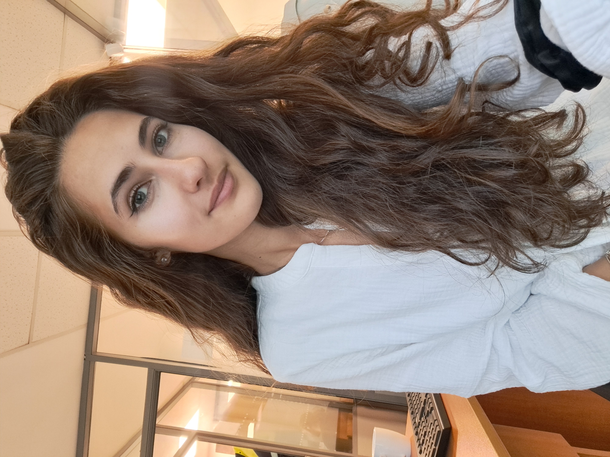 A young woman with long, wavy brown hair is indoors, possibly in an office setting. She is wearing a white shirt. A desk with a computer keyboard and a white cup is visible. Part of a window frame or room divider is in the background. The precise location within Sofia, Bulgaria cannot be determined from the visual data provided.