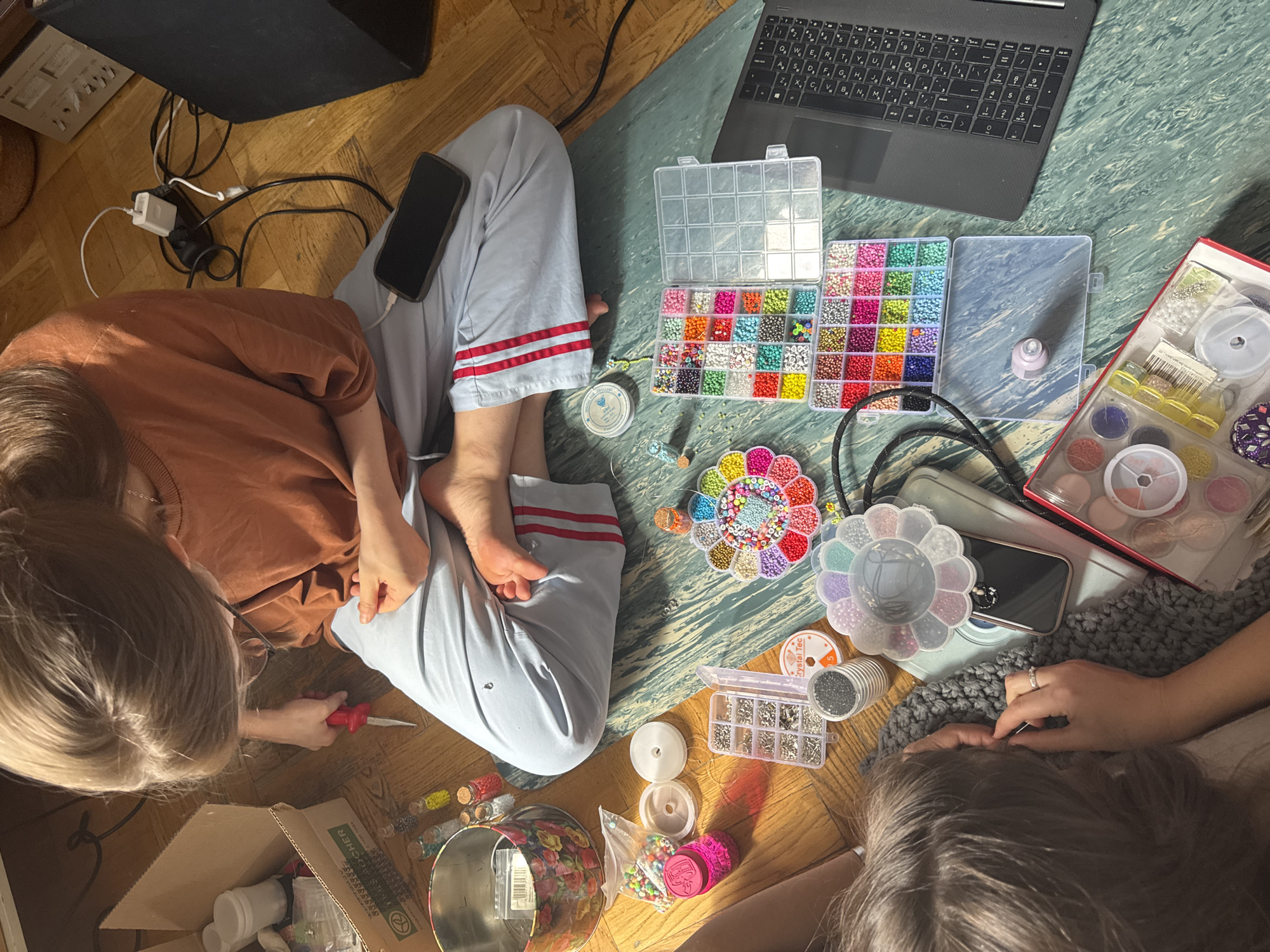 Two young people are seated on the floor amidst various crafting supplies. One person, wearing a brown shirt and light blue pants with red stripes, is using a tool with a red handle. A phone is visible in their lap. The other person, partially visible, is working with an unseen object, with their hands and arm visible. Scattered on the floor are containers of beads, spools of thread, small tools, a laptop, and a cardboard box. The scene takes place indoors on a wooden floor, covered in sections with carpet and possible crafting material. It is not possible to identify if this is in Moscow or Russia.