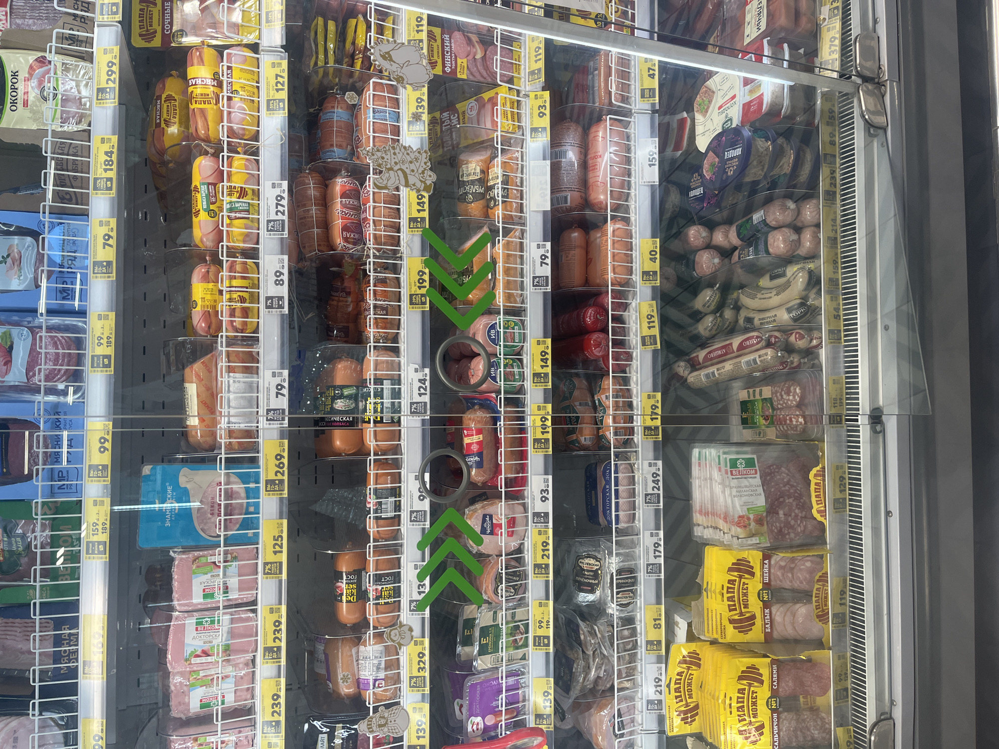 The image shows a refrigerated display case in a grocery store, likely a supermarket in Moscow, Russia. The case is filled with various processed meat products, predominantly sausages and ham. The products are packaged in plastic wrappers and some are displayed in bulk. Price tags are visible on most items, indicating different brands and varieties. The lighting is bright and reflects off the glass of the display case. There are no people visible in the image. The overall environment suggests a retail setting focused on the sale of perishable food items.