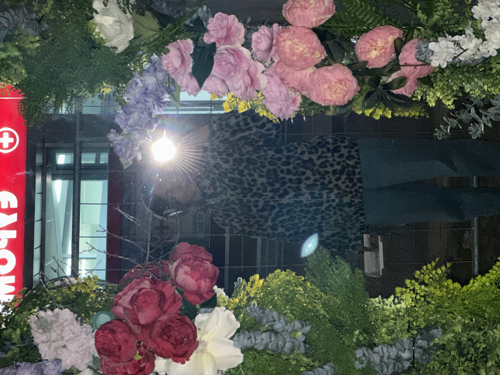 The image captures an upside-down view of a flower arrangement, predominantly featuring pink and red roses, white flowers, and various types of greenery, possibly artificial. The backdrop reveals a street scene, including a red sign with white text (likely advertising), a building with visible windows and metal bars, and a dark, leopard-print pattern above. The scene is lit by a bright light source, creating a starburst effect. The location appears to be Novi Sad, Serbia, based on the text on the window of the building.