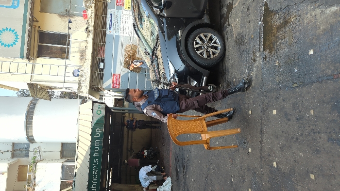 A police officer in uniform holds a rifle while carrying a tan plastic chair. He is standing on a street, next to a black car. Behind him is a garage or repair shop with people working inside. The building has signs for "lubricants from bp" and other businesses. The ground is dark and appears to be wet or oily.