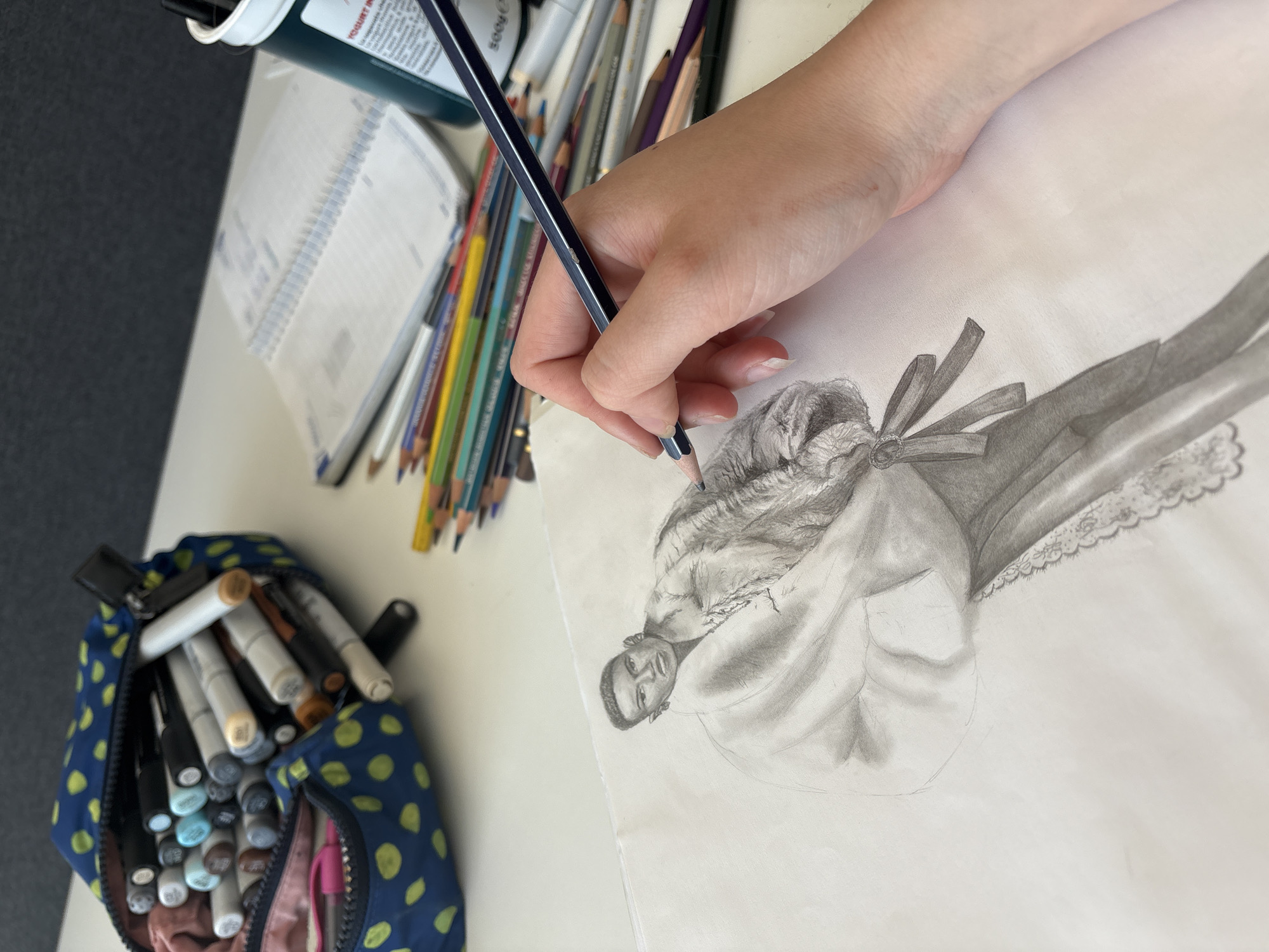 A person is drawing a pencil sketch of a historical figure on a white sheet of paper. The drawing depicts a dark-skinned man with short hair, wearing what appears to be a ruffled collar or lace detailing around the neck. The person's right hand is holding a pencil, poised over the drawing. The environment appears to be a desk or table with various art supplies. These include a pouch filled with markers, a can or container with brushes or pencils inside, a notepad, and a collection of colored pencils. There is nothing in the image to suggest that this is in the city of Milan, Italy.