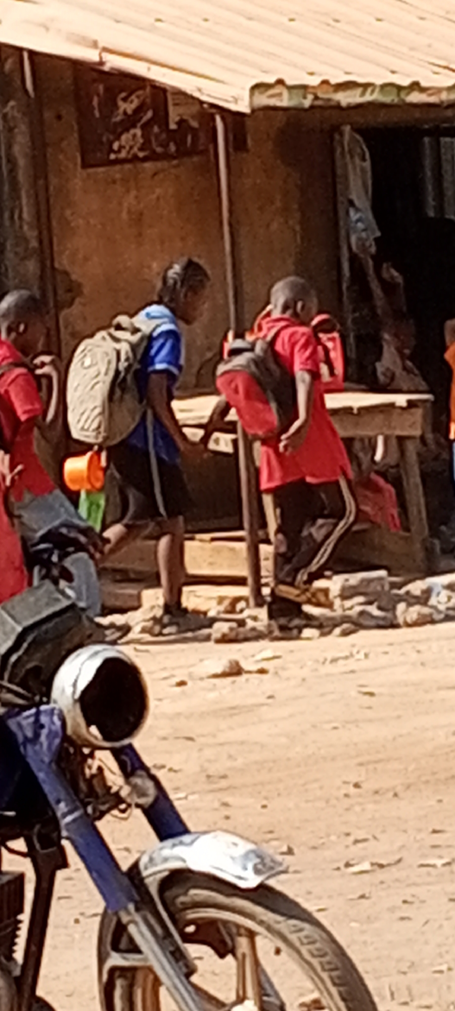 The image captures a street scene in Sabon Tasha, Nigeria. Three children wearing backpacks and school uniforms are walking along a dirt road past a building. One child is wearing a blue shirt, the other two red. A blue motorbike is parked in the foreground. The building has a corrugated metal roof and a dark interior. An individual sits inside the building.