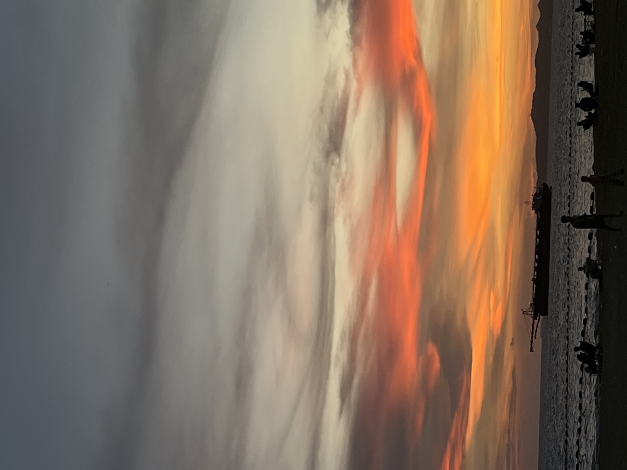 The image depicts a vibrant sunset over the water in Pattaya City, Thailand. Silhouetted figures of people are visible along the shoreline. A large ship or freighter is anchored offshore, with a crane visible on the water. The sky is filled with colorful clouds ranging from gray and white to orange and red hues. Mountains can be seen in the distance.