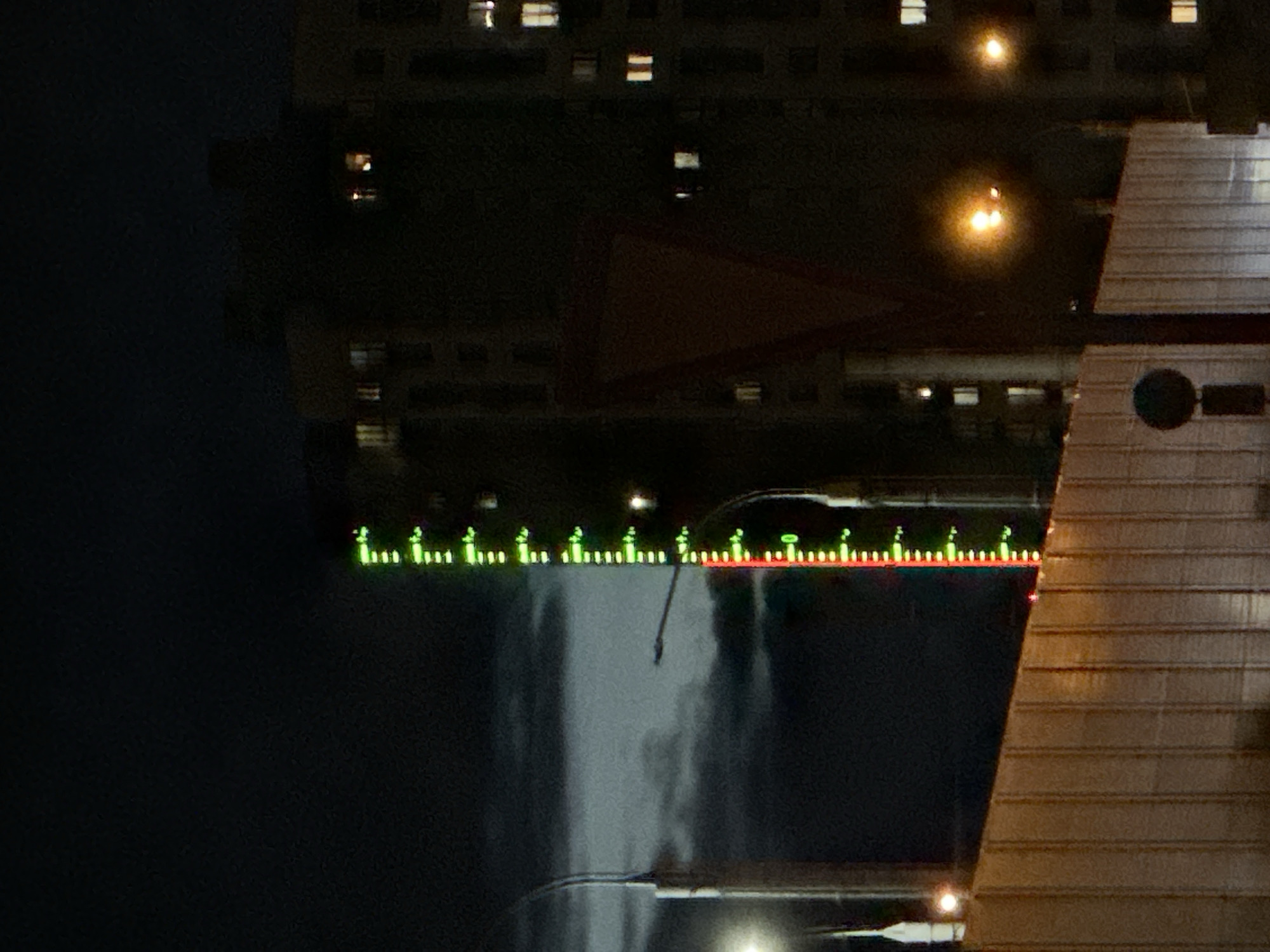 The image depicts a nighttime scene in Minsk, Belarus, showing the Svislach River. The dark water reflects the illuminated building and the sky above, creating an upside-down mirroring effect. A line of green and red lights, likely a decorative feature or a part of infrastructure, is visible along the water's edge. A portion of a building or structure with a circular window and horizontally layered panels is situated on the right side of the image, contributing to the overall composition of the cityscape. The time of day, lighting conditions, and the reflection on the river surface contribute to the scene's ambiance. The architecture and light colors help to identify the city as Minsk.