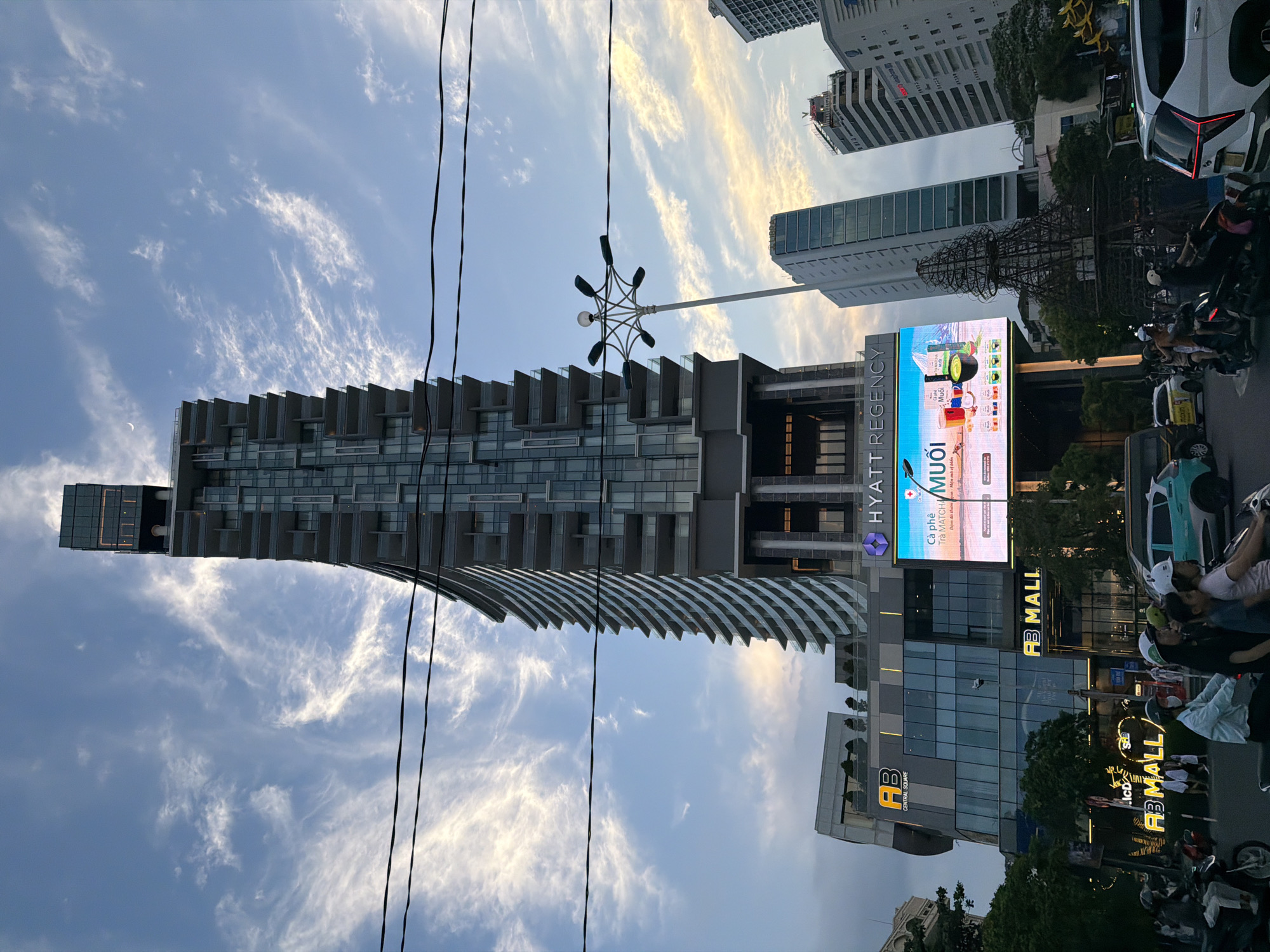 This is a vertical, low-angle shot of a street scene in Nha Trang Ward, Vietnam, likely in the late afternoon or early evening. The sky is a mix of blue with wispy, sunset-colored clouds. The foreground is dominated by power lines, and a modern, multi-story building with a curved, wavy facade stretches upwards. This building appears to be a hotel or luxury apartment complex, with many balconies.
To the right of this main building is a brightly lit digital billboard advertising "HYATT REGENCY" and "MUỐI" (which translates to salt), featuring images of coffee and tea. Below this, another building features prominent signage for "AB MALL" and "AB CENTRAL SQUARE". The street level is bustling with activity. A white car with distinctive red taillights is parked on the right, and several motorcycles with riders wearing helmets are visible, suggesting typical urban traffic. A few pedestrians are also present. The overall impression is a modern, vibrant urban environment in Vietnam.