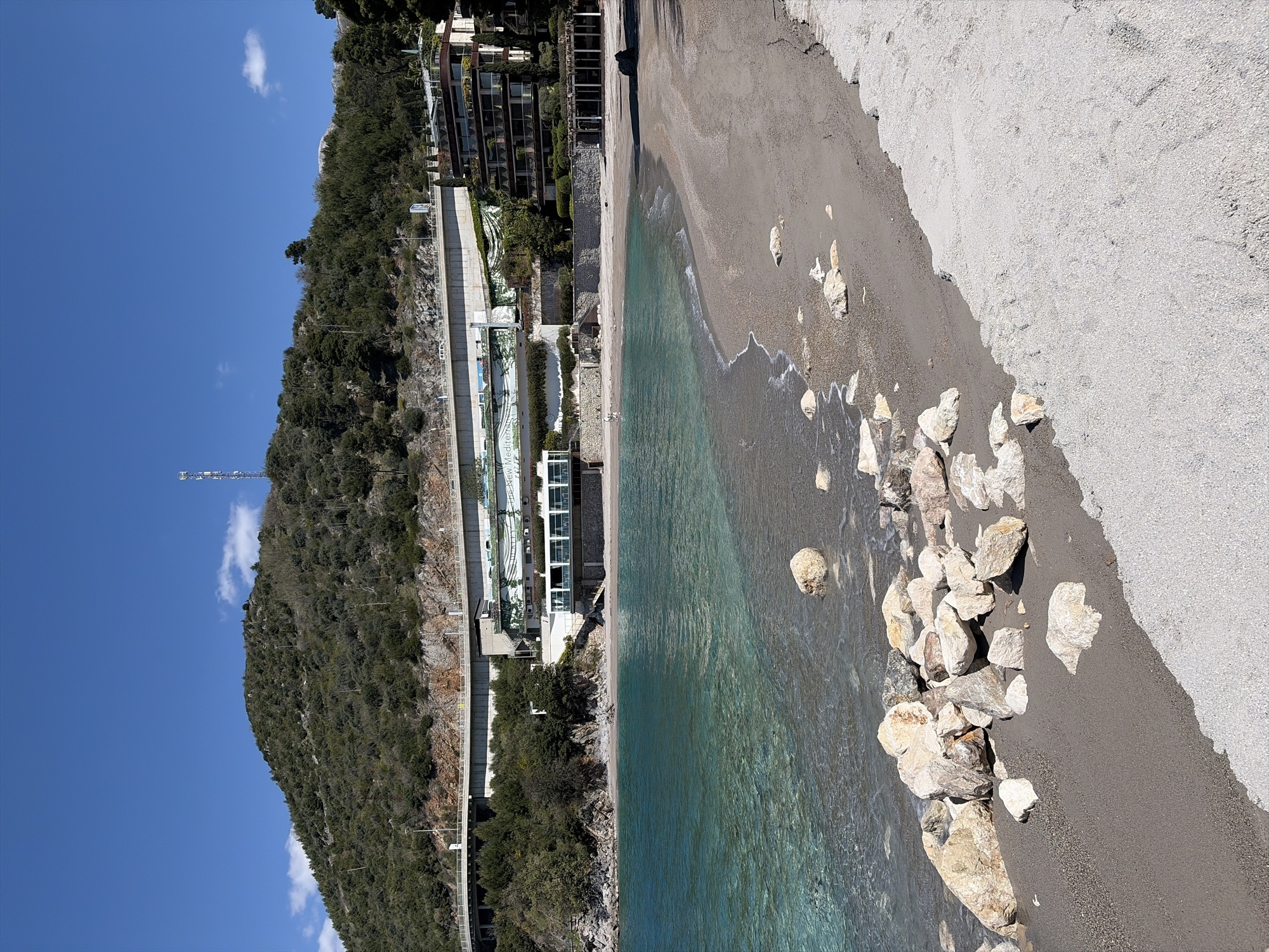 A high-angle view of a beach along the French Riviera on a sunny day reveals a scene of natural beauty juxtaposed with built infrastructure. The water is transparently blue and meets a dark sand beach that transitions into a light grey sand. A cluster of rocks sits at the water's edge, marking the boundary between the sea and land.
Along the coastline, structures are built into the hillside. A retaining wall supports the architecture, including modern buildings with glass facades. Lush green vegetation covers the slope above the structures, giving way to a rocky mountain. The sky above is clear blue, with a few scattered clouds. A cellular communication tower is seen rising from the top of the mountain. The location appears to be in the city of Cap-d'Ail, France.