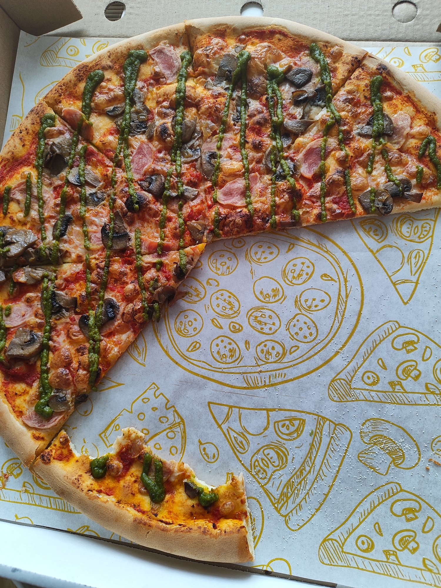 A partially consumed round pizza is presented within an open corrugated cardboard delivery box, viewed from a slightly elevated angle. Approximately two-thirds of the pizza remains, with a significant portion removed, forming a crescent shape, and one separate slice showing a bite mark. The pizza features a golden-brown crust and is topped with melted, bubbly cheese, sliced dark mushrooms, and thinly cut light pink meat, consistent with ham or bacon. A vibrant green, viscous sauce, resembling pesto, is drizzled in multiple wavy lines across the entire surface of the pizza. The interior of the pizza box is lined with white paper adorned with yellow outline illustrations of whole pizzas (one prominent central design featuring small circles, and other smaller designs), individual pizza slices, and mushroom caps. Scattered white granular material, consistent with flour or cornmeal, is visible on the box liner. A circular ventilation hole is present in the top-left visible section of the cardboard box lid. The pizza box rests on an undifferentiated white surface. No people are visible. The location of Marusino, Russia, cannot be determined from the visual information provided in the image.