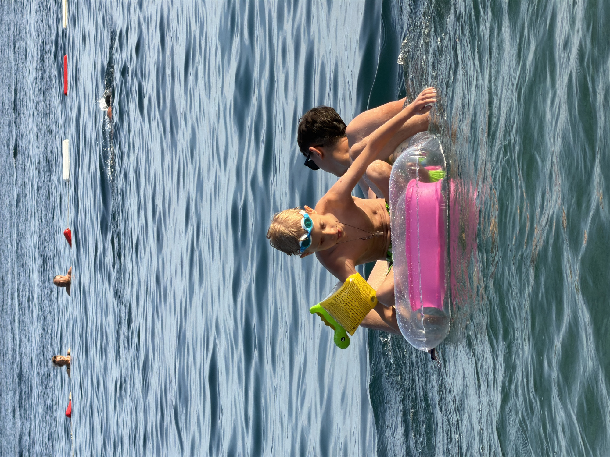 In the calm, blue waters of the Adriatic Sea, off the coast of Budva, Montenegro, two young boys are enjoying a day of swimming. The boy in the foreground, wearing blue goggles and a necklace, sits on a clear inflatable tube with a pink inner tube. He holds a yellow toy with a green alligator attached. Beside him, a second boy, wearing sunglasses and with his back to the camera, leans against him, his arm around the first boy. Both boys appear relaxed and are fully immersed in the water, their legs submerged. In the background, several other swimmers can be seen in the distance, some with buoys, indicating a popular recreational area. The sun glints off the water's surface, creating a serene and idyllic scene.