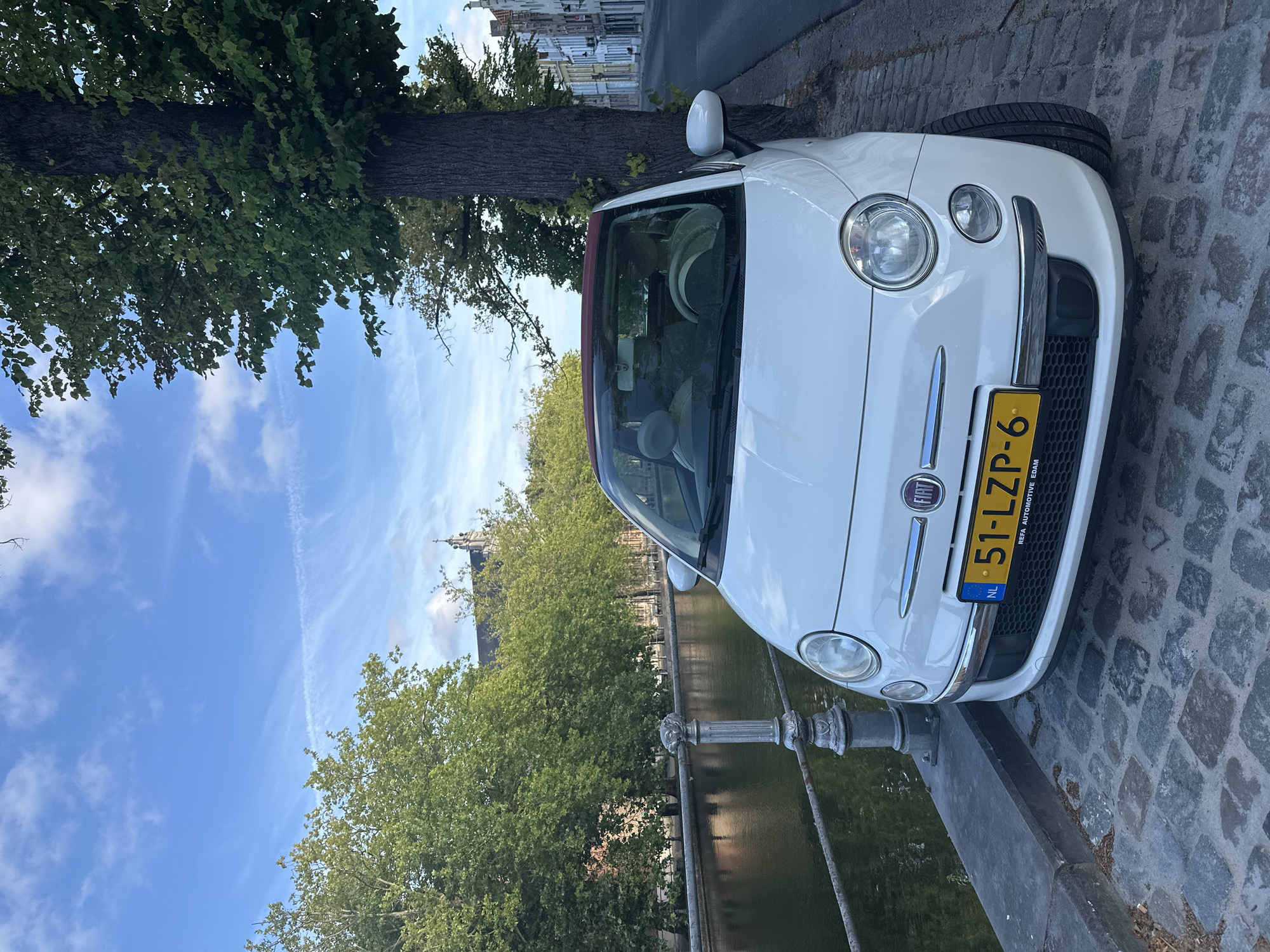 A white Fiat 500 convertible is parked on a cobblestone street next to a canal in Bruges, Belgium. The car, with its red soft top partially down, faces the viewer and is positioned on the right side of the frame. The license plate is visible, reading "51-LZP-6". Behind the car, a large tree with dark bark and green foliage dominates the right side of the image. The canal, reflecting the sky and trees, runs along the left side of the street, bordered by a stone embankment and an ornate metal railing. Across the canal, a line of lush green trees forms a backdrop, with a church spire visible in the distance. The street continues to the right, lined with historic buildings with colorful facades, indicative of Bruges' architectural style. The sky is blue with scattered white clouds and a faint contrail. No people are visible in the immediate vicinity of the car.