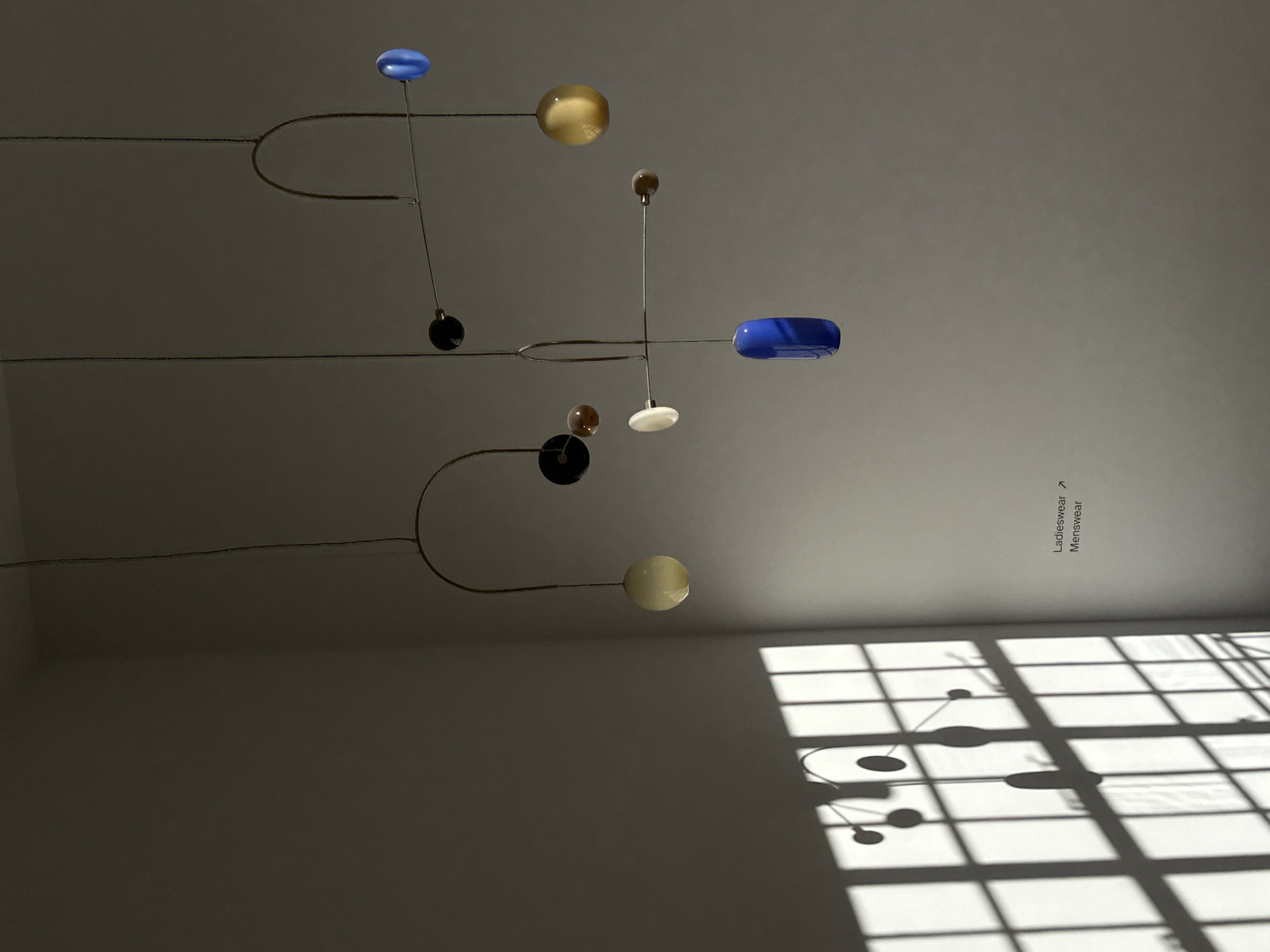This image depicts a minimalist indoor scene, likely a modern retail or gallery space in London, United Kingdom. A sophisticated mobile-like lighting fixture is suspended from a light-colored ceiling, serving as the central object. This fixture comprises thin metallic rods that balance several spherical and capsule-shaped elements in various sizes and colors, including vibrant blue, golden-yellow, dark brown/black, and off-white.
Strong, angular shadows from a large, gridded window are cast across the lower right wall, indicating direct sunlight and a sunny daytime, likely morning or late afternoon. The mobile itself also casts abstract shadows onto the wall, interacting with the window shadows to create a complex geometric pattern. The room's walls and ceiling are a uniform light grey or off-white.
On the plain wall, vertically oriented text serves as a directional sign: "Ladieswear" with an upward arrow, and beneath it, "Menswear." This text strongly suggests a commercial, possibly a department store or high-end boutique setting. No people are present, allowing the focus to remain on the interplay of light, shadow, and modern design elements, creating a serene and artistic ambiance.
