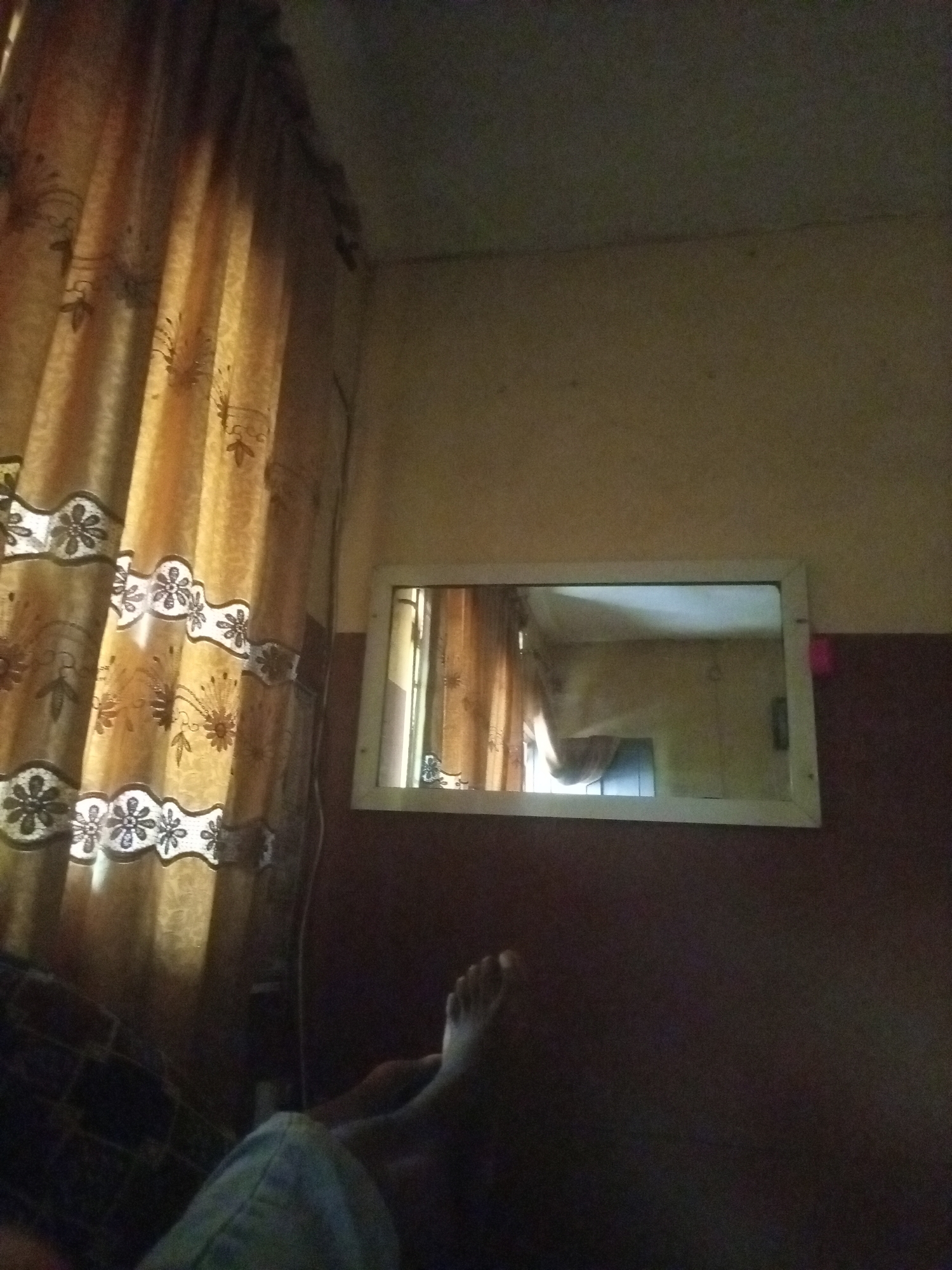 The image is a low-angle, indoor shot taken in what appears to be a bedroom in Onitsha, Nigeria. In the foreground, a person's bare feet and lower legs are visible, resting on a dark patterned surface that could be a bed or a chair. The person is wearing light-colored pants. To the left, a thick, golden-yellow curtain with a repeating floral pattern of white and black flowers is drawn, partially obscuring the background. The wall to the right is a muted beige color, transitioning to a darker brown lower down. Mounted on the wall is a rectangular mirror with a white frame. The mirror reflects the opposite side of the room, showing more of the golden curtain, a window with light streaming through, and a ceiling. The lighting in the room is dim, with light primarily coming from the unseen window. The overall impression is one of a simple, lived-in space.