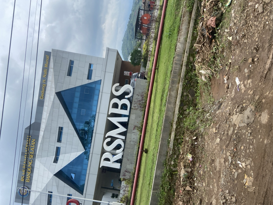 The image shows a view of the front entrance of RS Muhammadiyah Bandung Selatan, a hospital located at latitude -7.022 and longitude 107.68 in the city of Ciparay, Indonesia. The hospital building is a modern structure with a geometric facade featuring large glass windows. The building has a sign that reads "Gedung H. Nafli Munaf," which likely indicates the name of the building. A large sign in front of the building displays the hospital's name, "RSMBS," in bold, white lettering. In the foreground, a red pipe runs across a patch of grass, bordered by a concrete wall. The ground is covered in dirt and debris, including small rocks, twigs, and plastic bags. The hospital is located on a sloping lot, with a small hill in the background. The sky is partly cloudy.