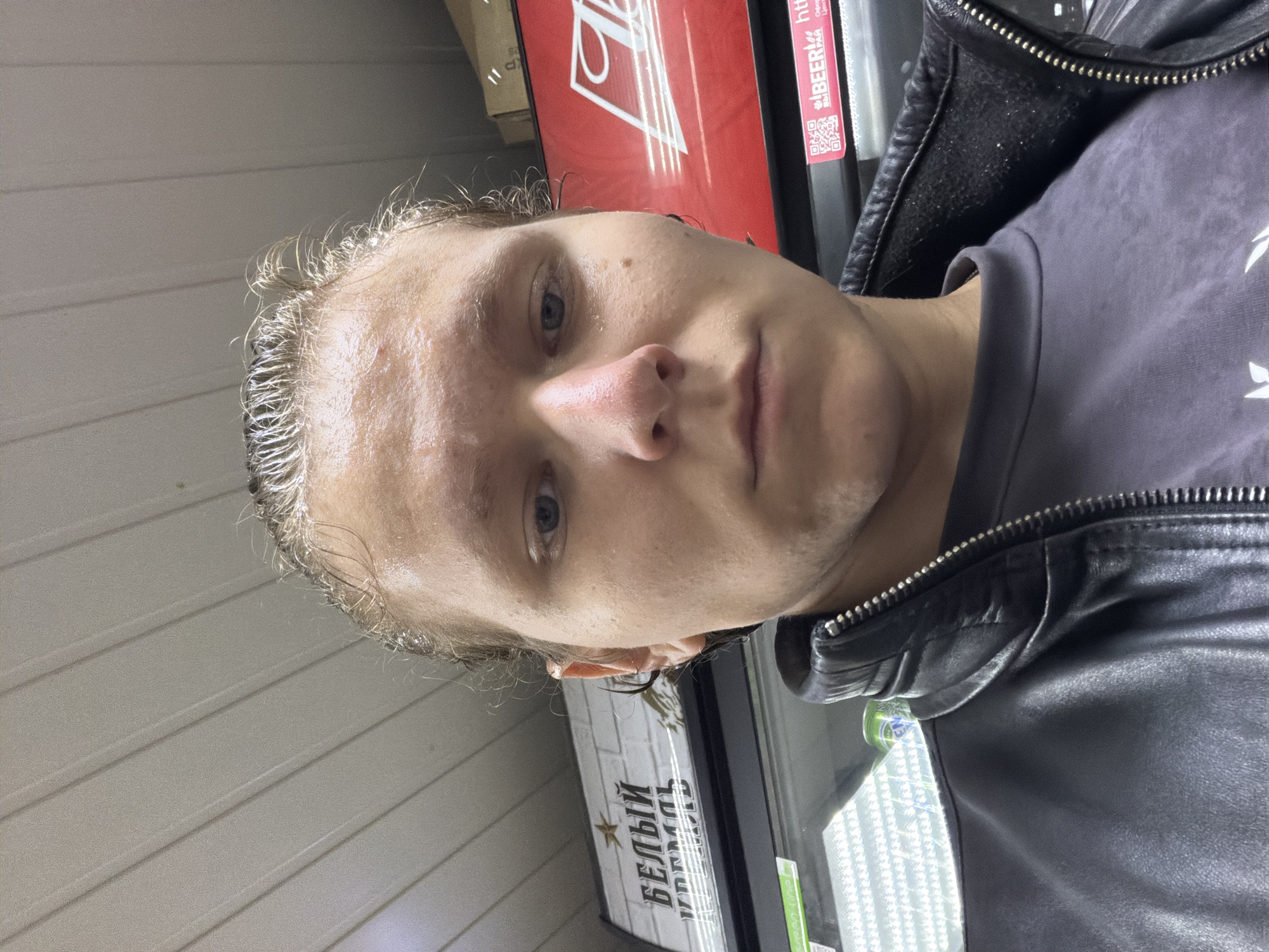 A close-up, low-angle selfie features a young adult male with wet, slicked-back blonde hair and a dark leather jacket. He appears to be in a brightly lit, indoor commercial setting, possibly a convenience store or market, given the presence of refrigerated displays with visible products and signage in the background. One sign displays text in Russian, including "БЕЛЫЙ КРЕМЛЬ," and another features a red logo with a white outline that resembles the Budweiser logo. The environment suggests an urban or semi-urban location, consistent with Chistopol, Russia. The subject's face is illuminated from above, highlighting sweat on his forehead and nose, suggesting exertion or humidity.